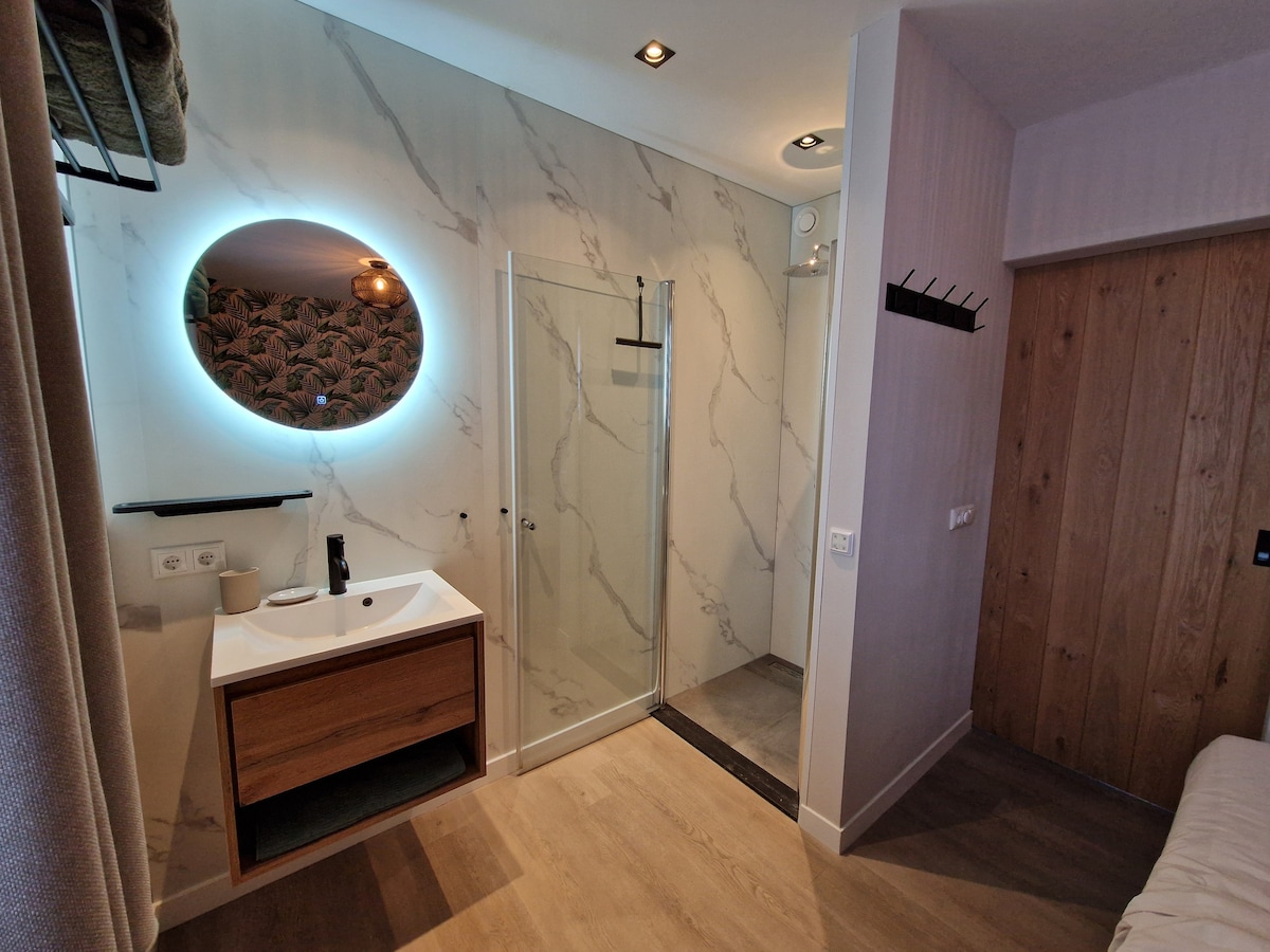 The modern bathroom features a minimalist design with a wooden vanity and a circular mirror framed by soft lighting. An in-wall shower is enclosed by glass, showcasing stylish marble tile accents. A towel rack is mounted on the wall, complementing the overall aesthetic.
