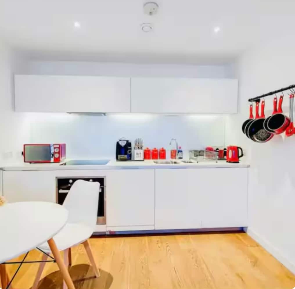 A modern kitchen is presented with sleek white cabinetry and a minimalist design. Essential appliances, including a microwave and coffee maker, are featured. A small round dining table with a white chair occupies the space. Cookware is neatly organized on a rack, adding to the kitchen's functionality.