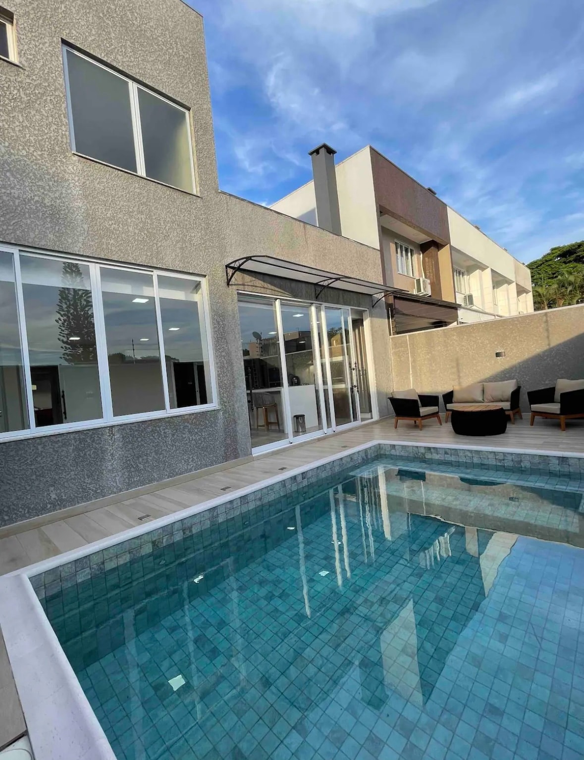 A heated pool is situated outside, surrounded by a wooden deck. Large glass doors provide access to the interior, creating a connection between indoor and outdoor spaces. Comfortable seating is arranged nearby, allowing for relaxation and enjoyment of the sun.
