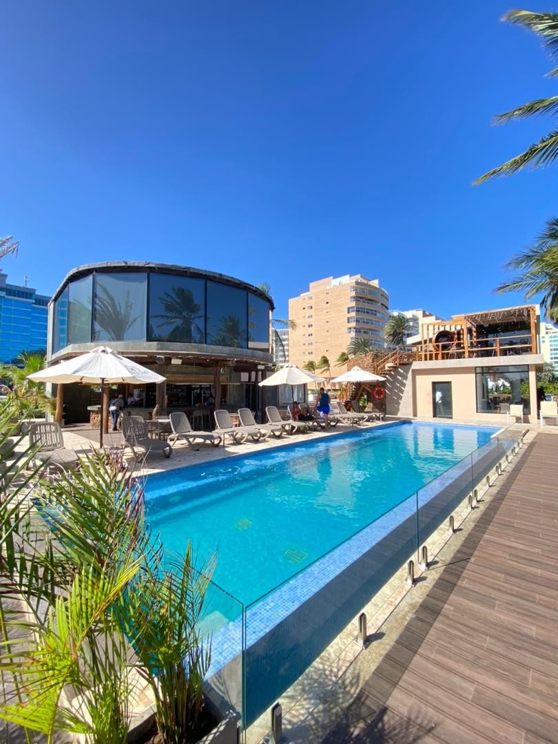 A modern outdoor pool area is surrounded by palm trees and lounge chairs beneath white umbrellas. A circular glass structure and multi-level building are visible in the background against a clear blue sky, creating a relaxed atmosphere for guests.