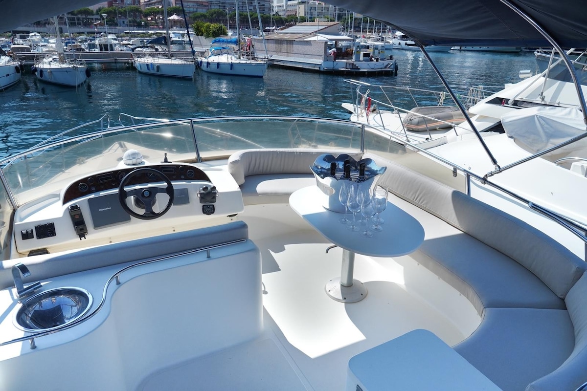 A spacious upper deck area is presented, featuring a circular table surrounded by comfortable seating. A steering wheel is positioned at the helm, offering a clear view of the marina. Glassware and a bottle are arranged elegantly on the table, with boats visible in the background.