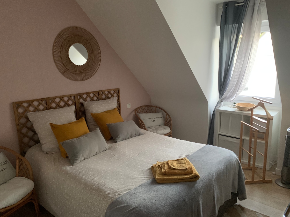 A light and airy bedroom featuring a spacious bed adorned with neutral bedding and decorative pillows in shades of yellow and gray. A circular mirror is positioned above the headboard, while natural light filters through the window, complemented by sheer and dark curtains.
