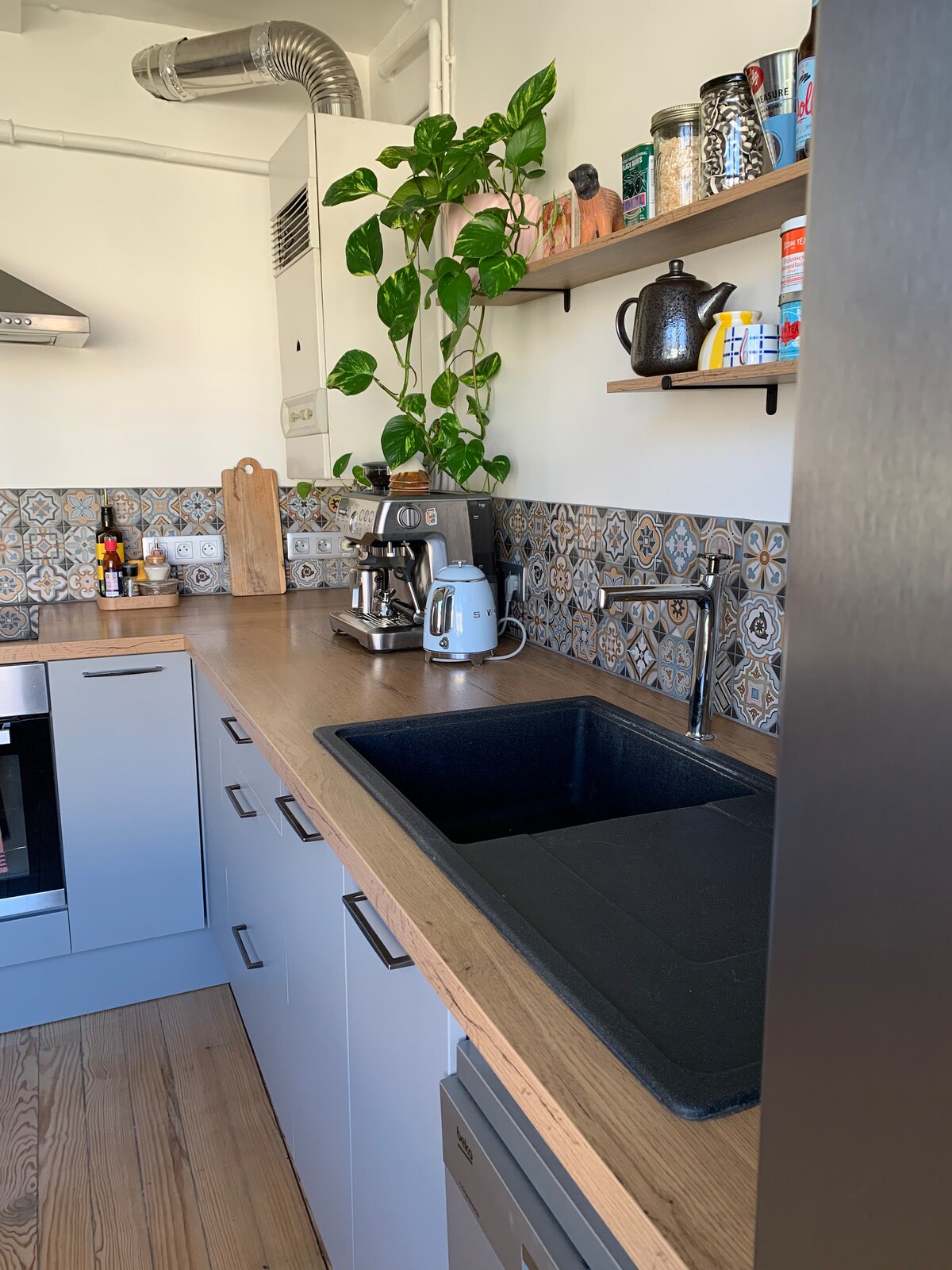 The kitchen features a sleek countertop with a black sink and modern appliances, including a coffee machine and kettle. Open shelving displays an assortment of jars and crockery, while a potted plant adds a touch of greenery to the space.