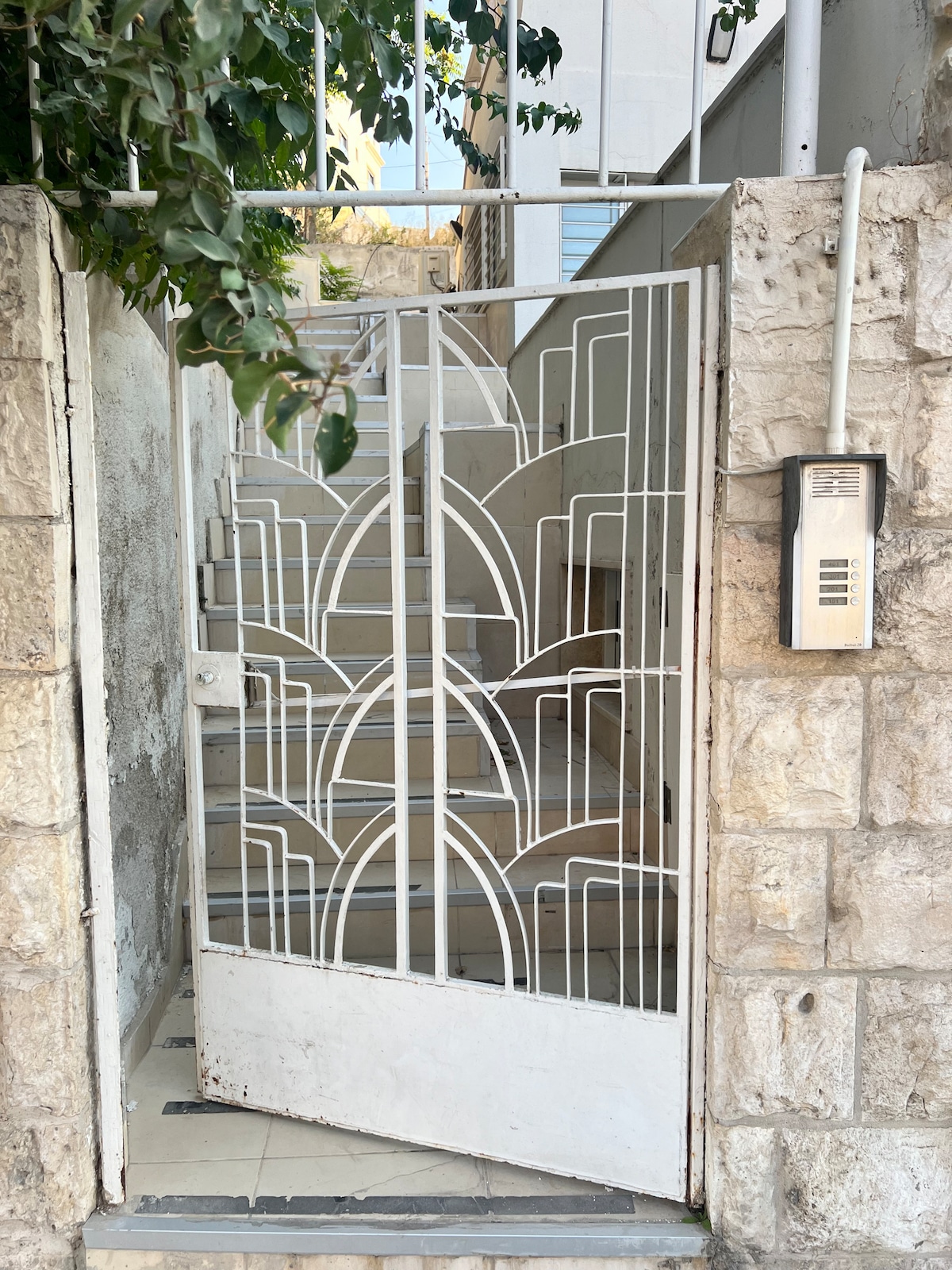 A decorative white metal gate features an artistic geometric design, leading to a staircase that ascends out of view. Surrounding greenery can be seen framing the entrance, which is flanked by a textured stone wall.