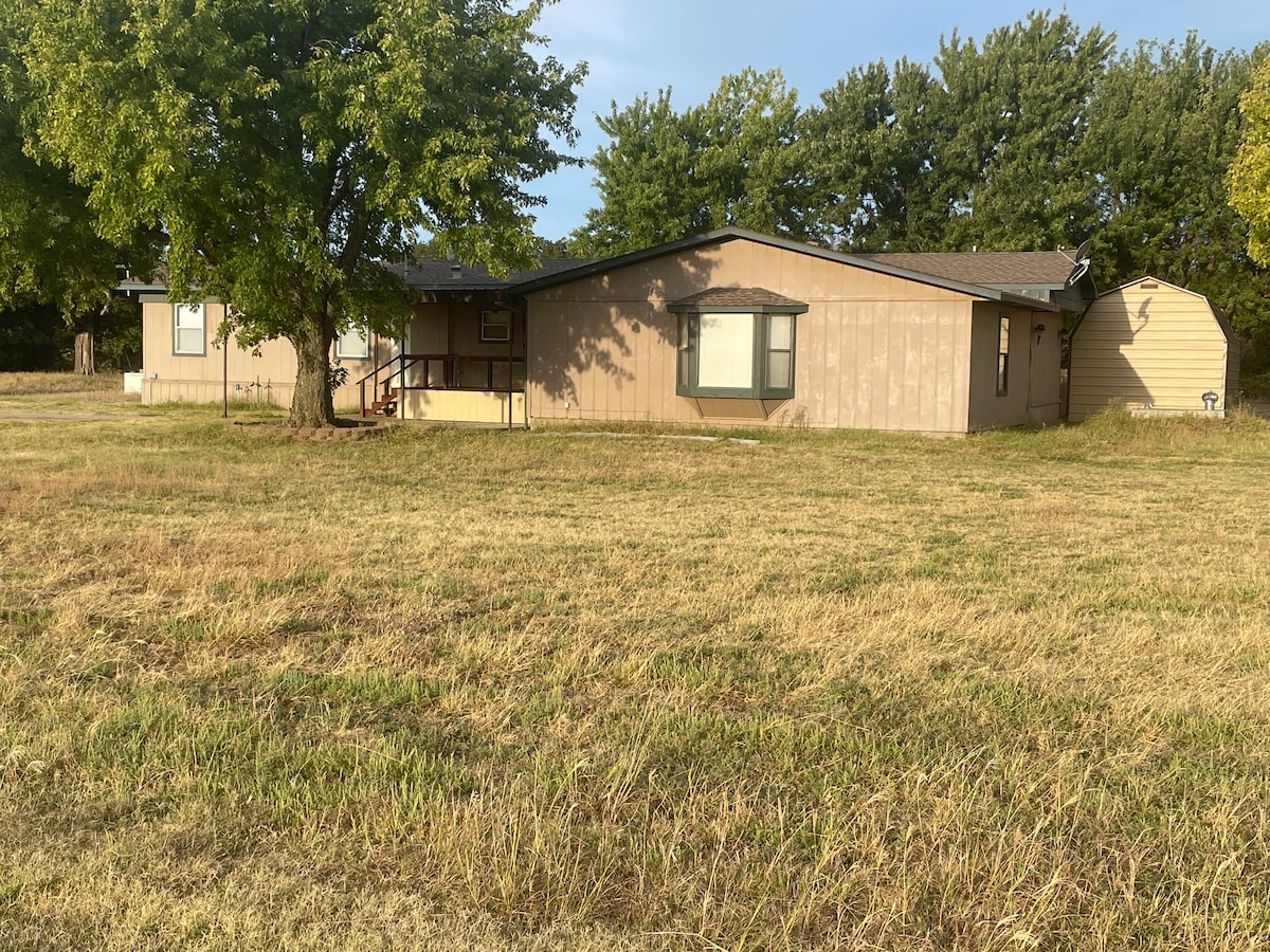 The exterior of a single-story building is shown, surrounded by a spacious grass lawn. Two large trees provide shade in the front yard, and the house features a wooden porch with steps leading to the entrance. Several windows are visible on the front facade.