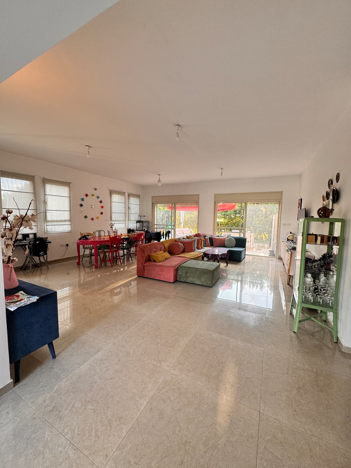 A spacious living area is filled with natural light from large windows. A colorful sectional sofa is arranged with coordinating pillows, accompanied by a modern dining table. Light-colored tiled flooring reflects the bright ambiance, enhancing the open layout that connects the indoor and outdoor spaces.