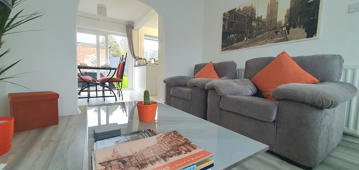A spacious living area is featured with comfortable gray sofas adorned with orange cushions. A glass coffee table sits in front, showcasing books and a small potted plant. An adjacent dining space is visible, with a table surrounded by chairs and bright natural light from large windows.