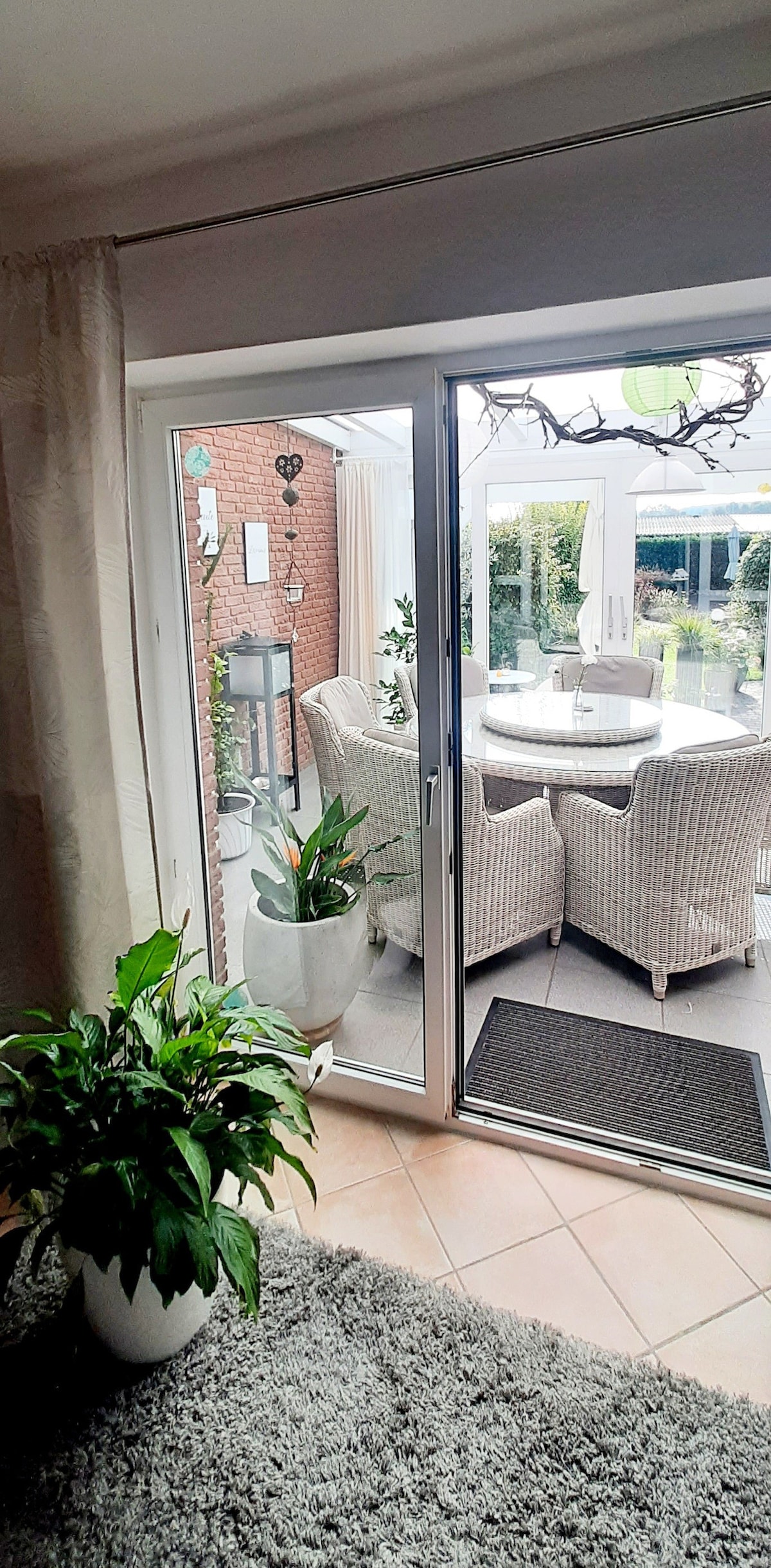 A spacious entrance opens to a winter garden through large glass doors. A round dining table is surrounded by rattan chairs, while potted plants add greenery to the space. Natural light creates a bright atmosphere, with a soft rug visible in the foreground.