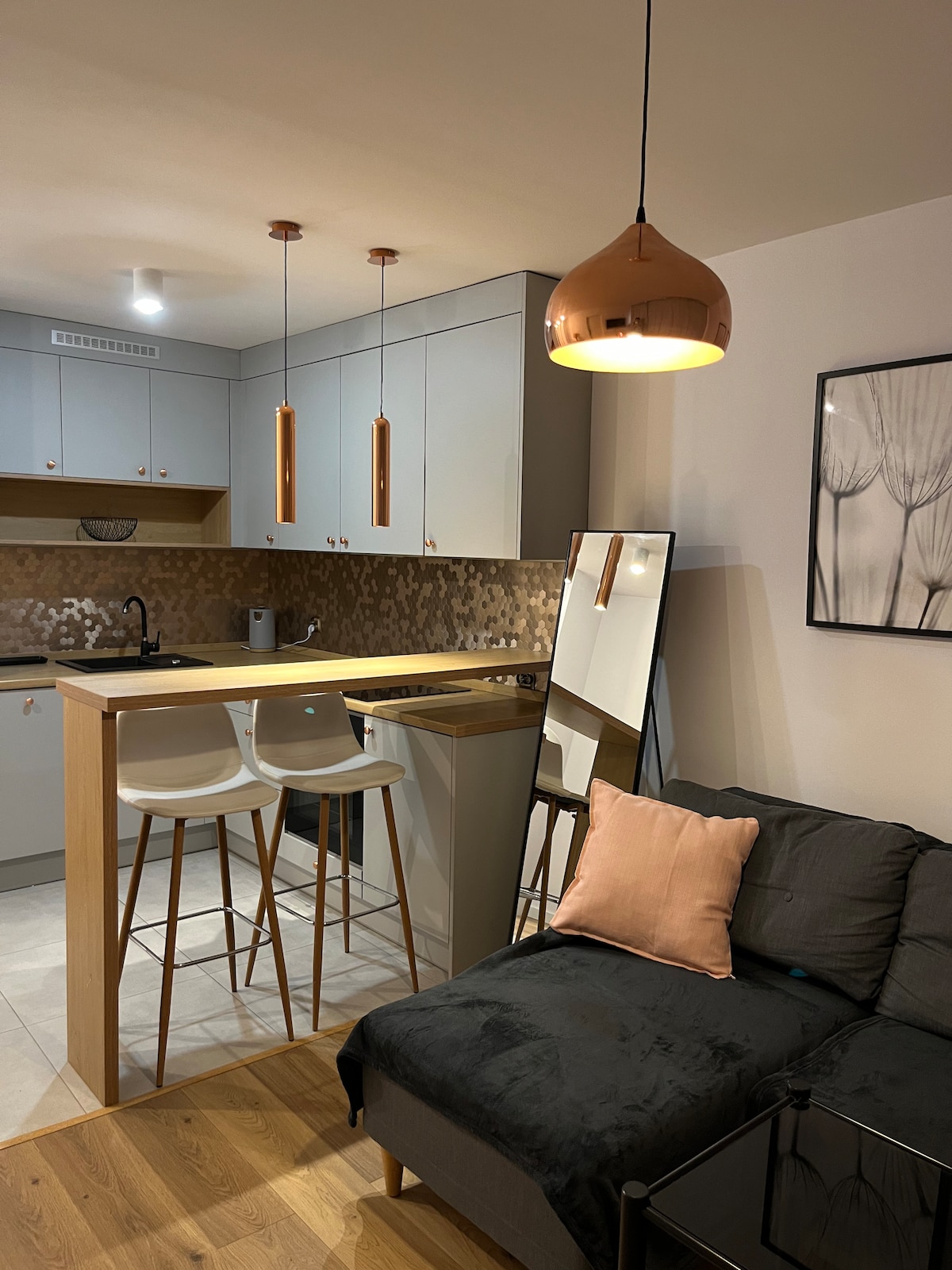 A modern living space features a sleek kitchenette with wood accents and stylish pendant lighting. Two upholstered stools are positioned at a small breakfast bar. A mirror leans against the wall next to a dark sofa adorned with a soft pink cushion, complemented by a minimalistic wall art piece.