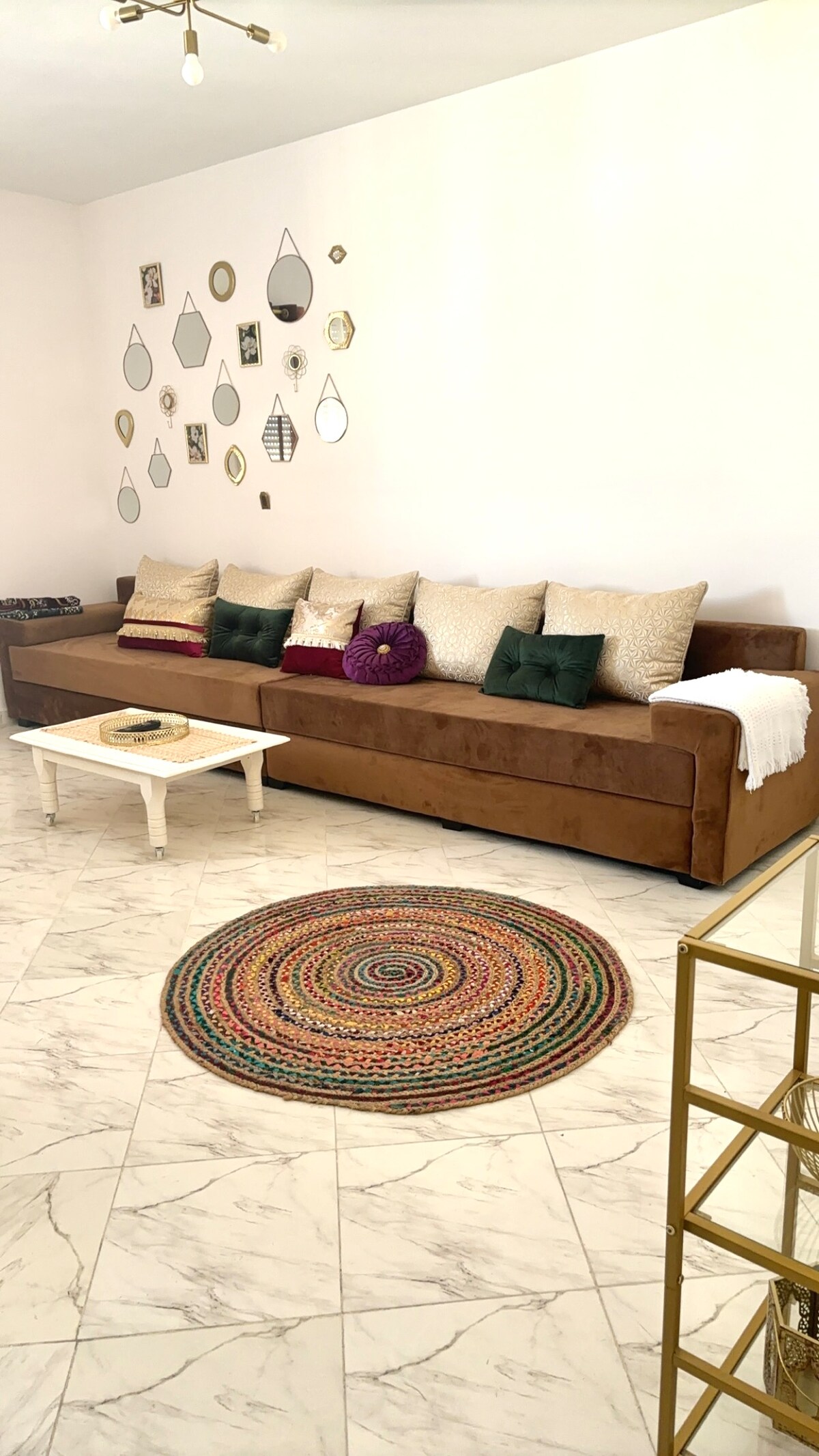 A spacious sitting area is highlighted by a large brown sofa adorned with various decorative cushions. A round, textured rug anchors the space, while a small white coffee table sits in front. Decorative mirrors are arranged on the wall, adding visual interest.