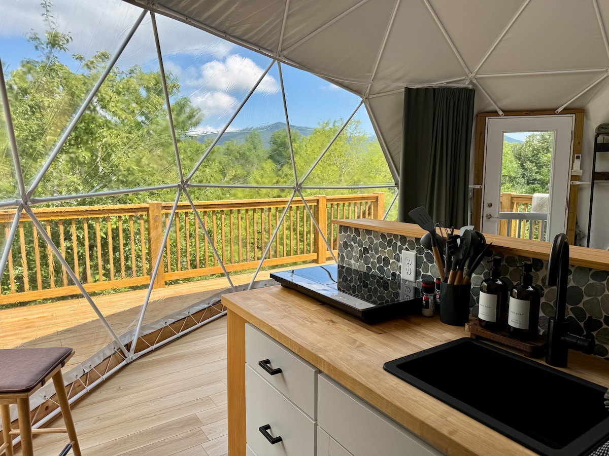 The interior of the glamping dome features a well-equipped kitchen with a wooden countertop and modern amenities. A spacious window offers unobstructed mountain views, while the wooden deck is visible through the transparent dome structure. A stool is positioned near the kitchen area.