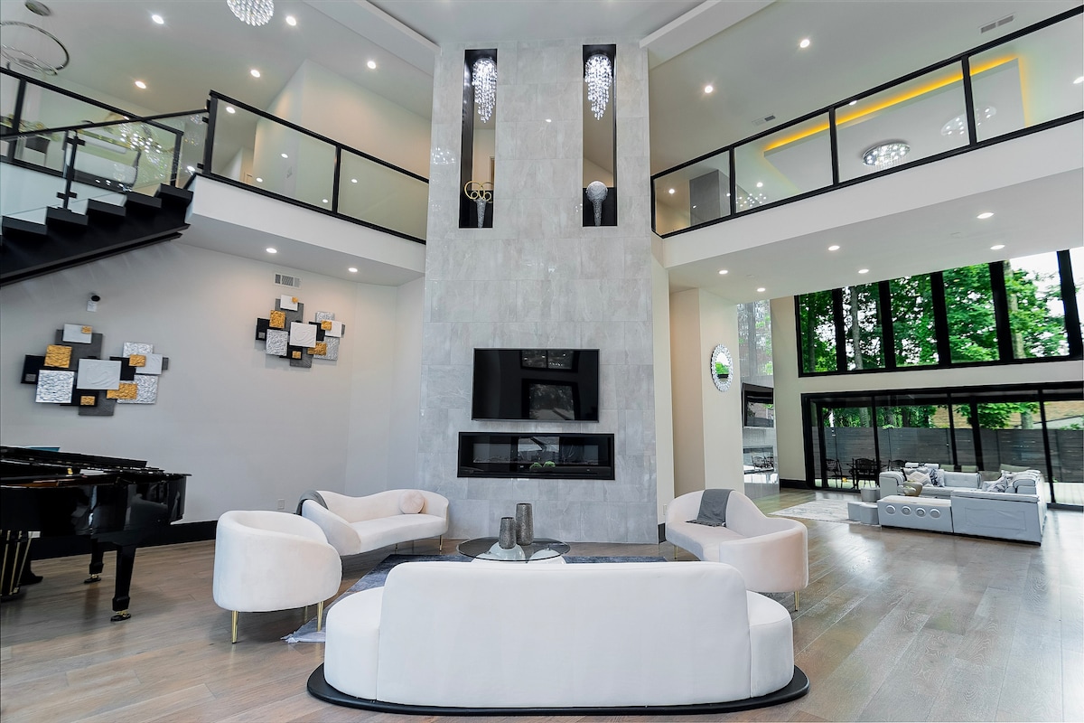 The expansive living area features high ceilings and large windows, allowing abundant natural light to fill the space. A modern fireplace is centered on a sleek wall, surrounded by elegant seating arrangements. Artistic wall decor and a grand piano enhance the sophisticated ambiance of the home.