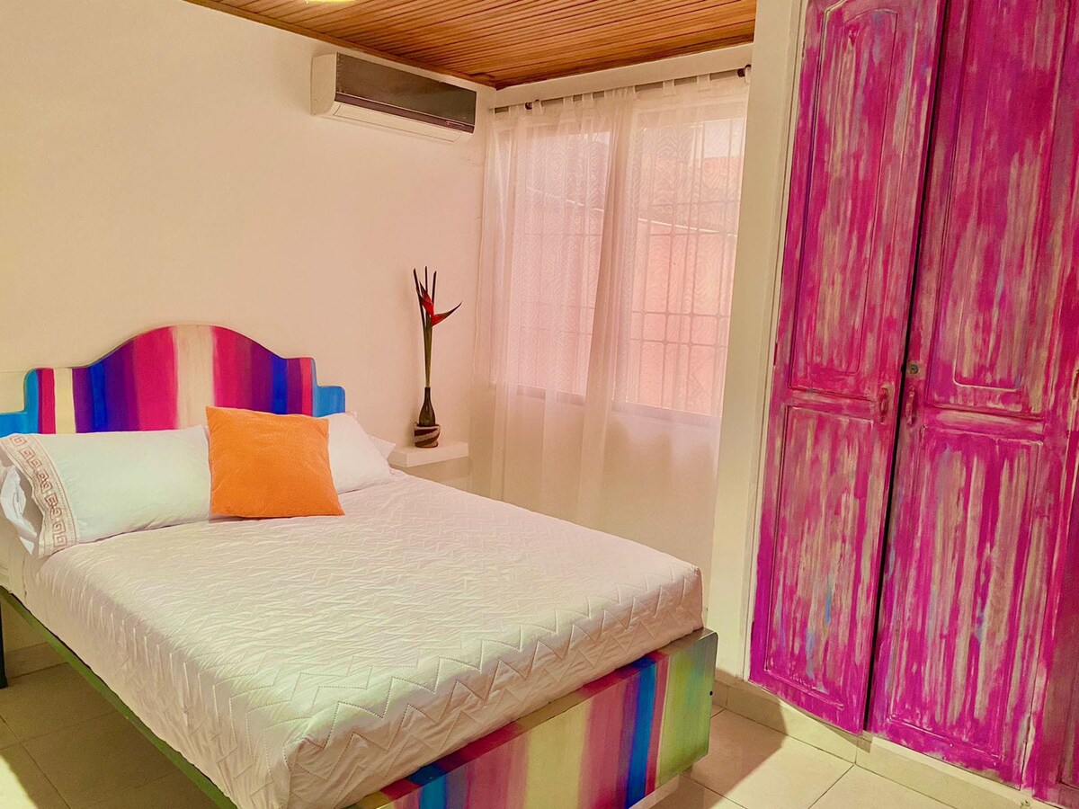 A vibrant sleeping area features a spacious bed with a striped headboard and an orange accent pillow. Natural light filters through sheer curtains covering a window, while a wooden cabinet painted in pink adds a pop of color to the room's decor.