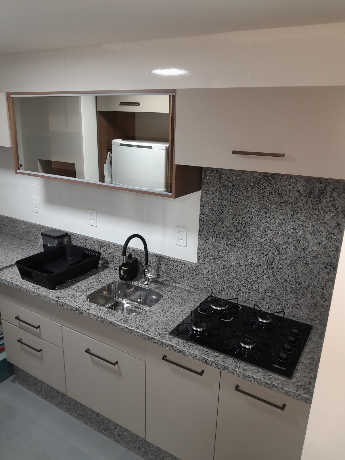 A modern kitchen area features an integrated granite countertop with a stainless steel sink and a gas stove. Above the countertop, a mirrored cabinet reflects the welcoming space. A refrigerator is visible in the background, with organized storage items neatly arranged on the counter.