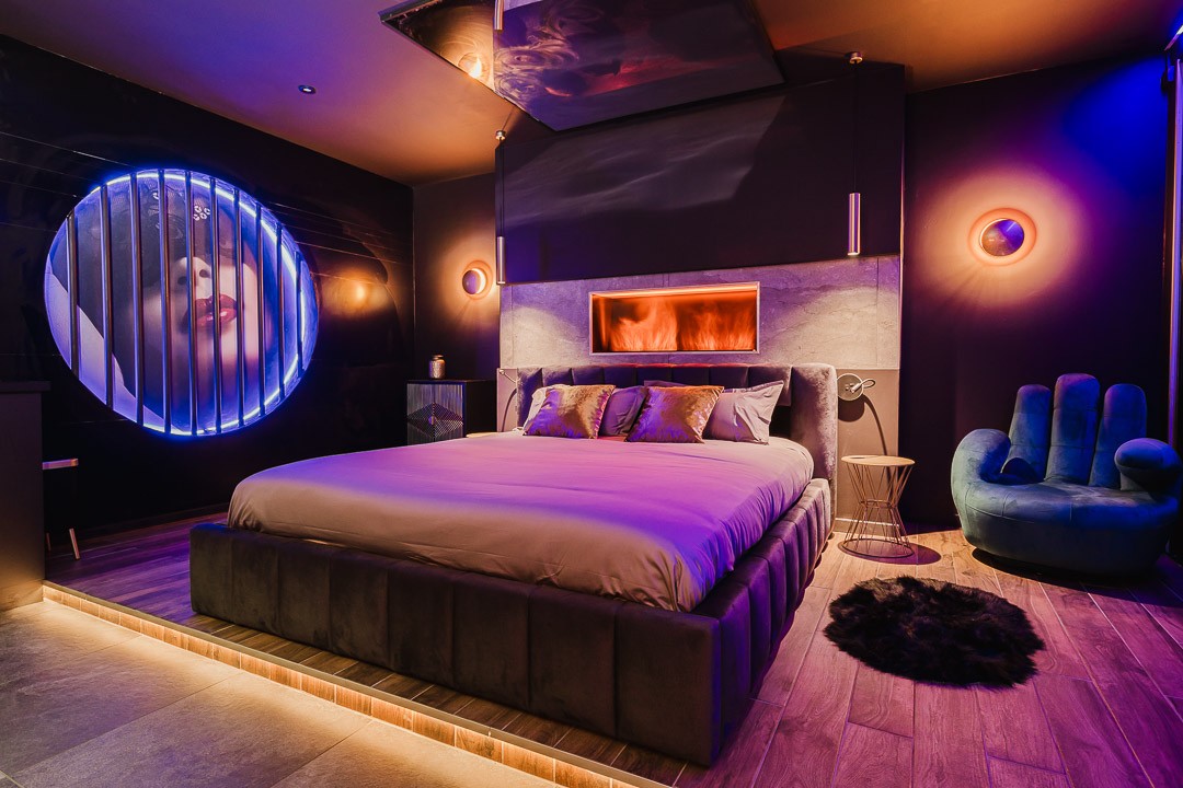 A spacious bed with plush pillows is centered in a dimly lit room, enhanced by colorful lighting. A modern electric fireplace provides warmth, while an artistic feature with a circular design adds visual interest. A stylish chair complements the contemporary decor.