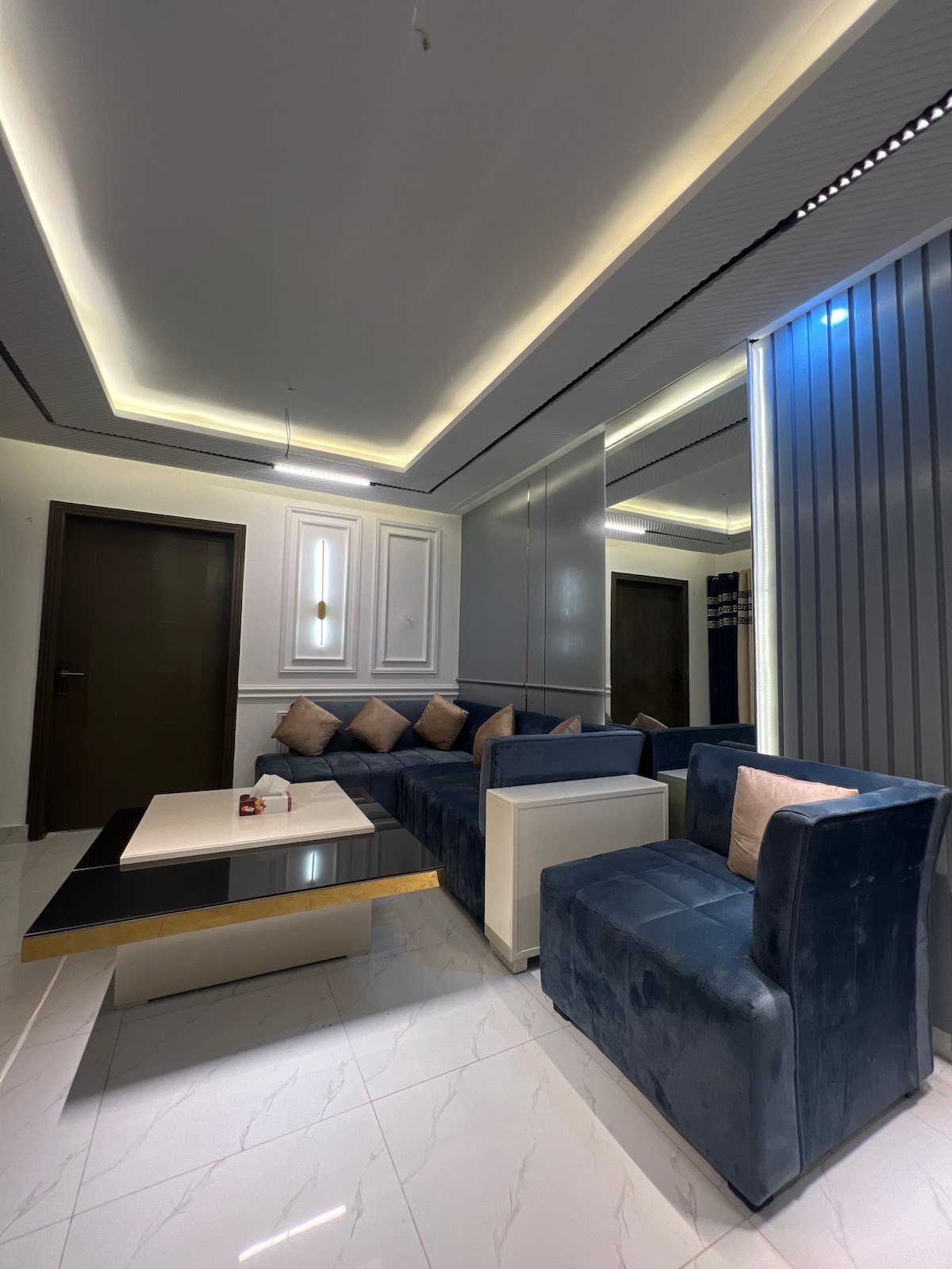 A modern living area is showcased, featuring a plush sectional sofa in deep blue with decorative cushions. A sleek coffee table occupies the center, surrounded by light-colored walls that reflect the ambient lighting, enhancing the spacious feel of the room.