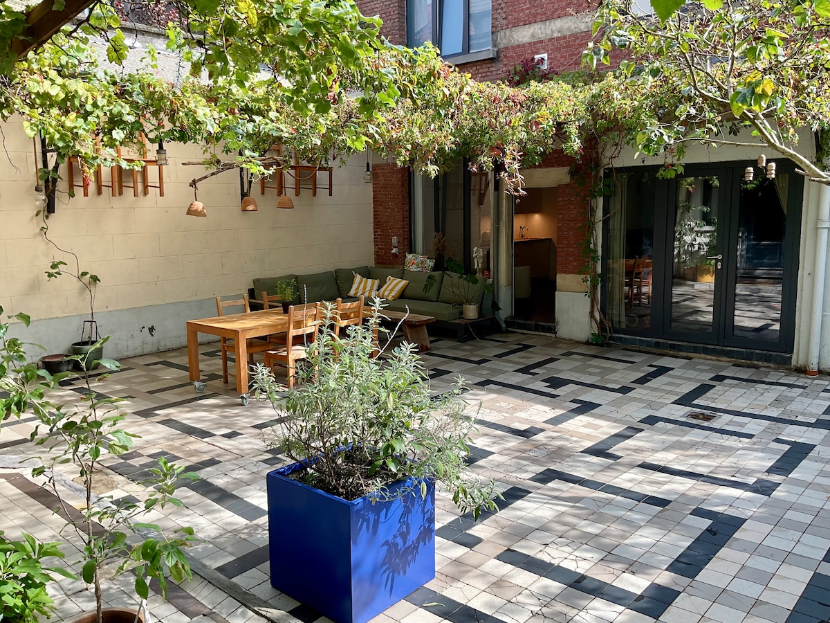 A communal courtyard is depicted, featuring a large wooden dining table surrounded by several chairs. A comfortable seating area is visible, adorned with cushions. Potted plants are scattered throughout, and a vibrant blue planter contains lush greenery underneath a canopy of climbing vines.