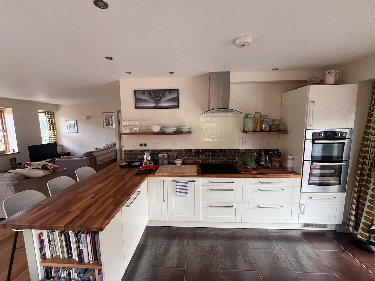 An equipped kitchen is displayed with a wooden countertop and modern appliances. White cabinetry complements the dark flooring, and a dining area with white chairs is visible nearby. Windows allow natural light to fill the space, with decorative items arranged on the shelves.
