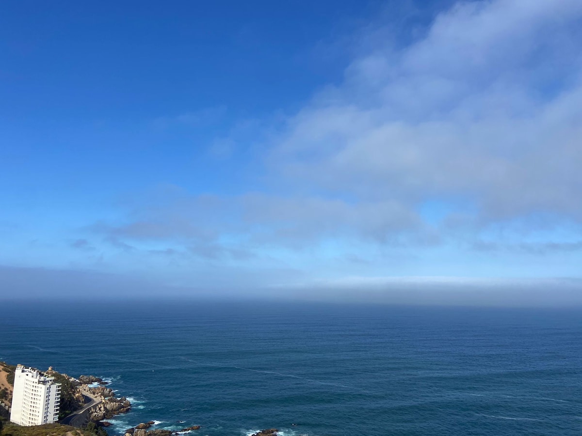 An expansive view of the ocean under a clear blue sky is presented, showcasing calm waters that stretch to the horizon. The coastline is visible in the foreground, with a white building nestled among the rocks, contributing to the scenic landscape.