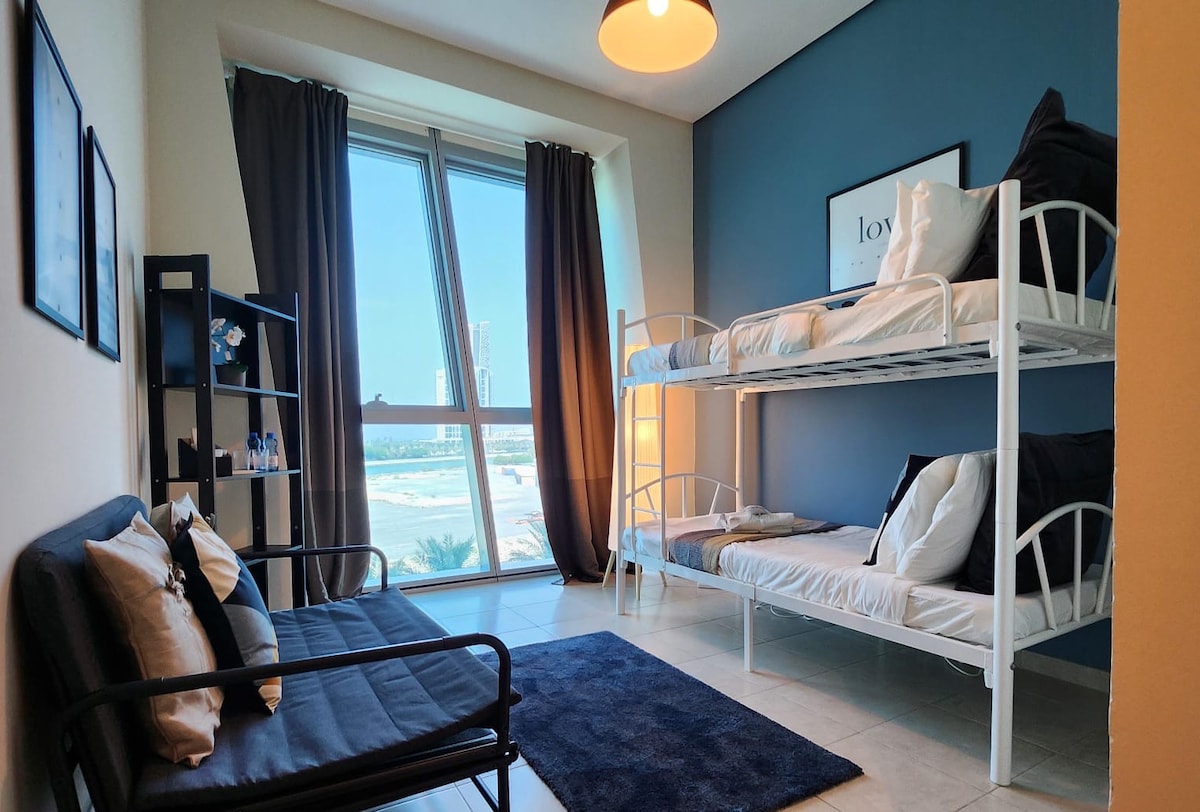 A bright bedroom features a bunk bed with two single beds and a convertible sofa. Large windows provide natural light and views of the outdoors. Neutral decor is complemented by dark curtains and a navy accent wall, enhancing the inviting space.