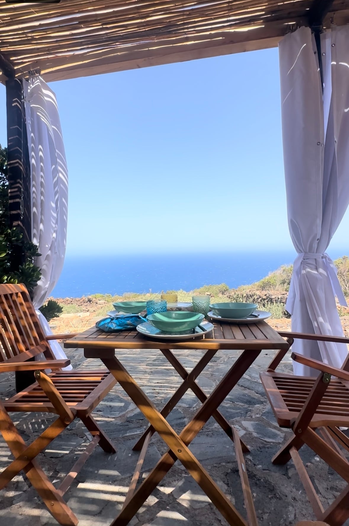 A small outdoor dining area is set for breakfast, featuring a wooden table with light blue and white dishware. Two folding chairs provide seating. The view beyond shows an expansive ocean under a clear blue sky, framed by sheer white curtains fluttering in the breeze.