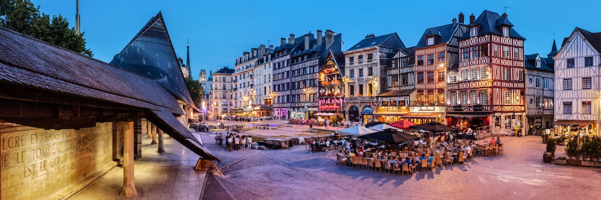 A vibrant square in Rouen’s old town is depicted at dusk, featuring charming half-timbered buildings and outdoor seating areas. Soft lighting from cafes creates an inviting atmosphere, and a cobblestone pathway leads through the scene, while decorative lights enhance the evening ambiance.