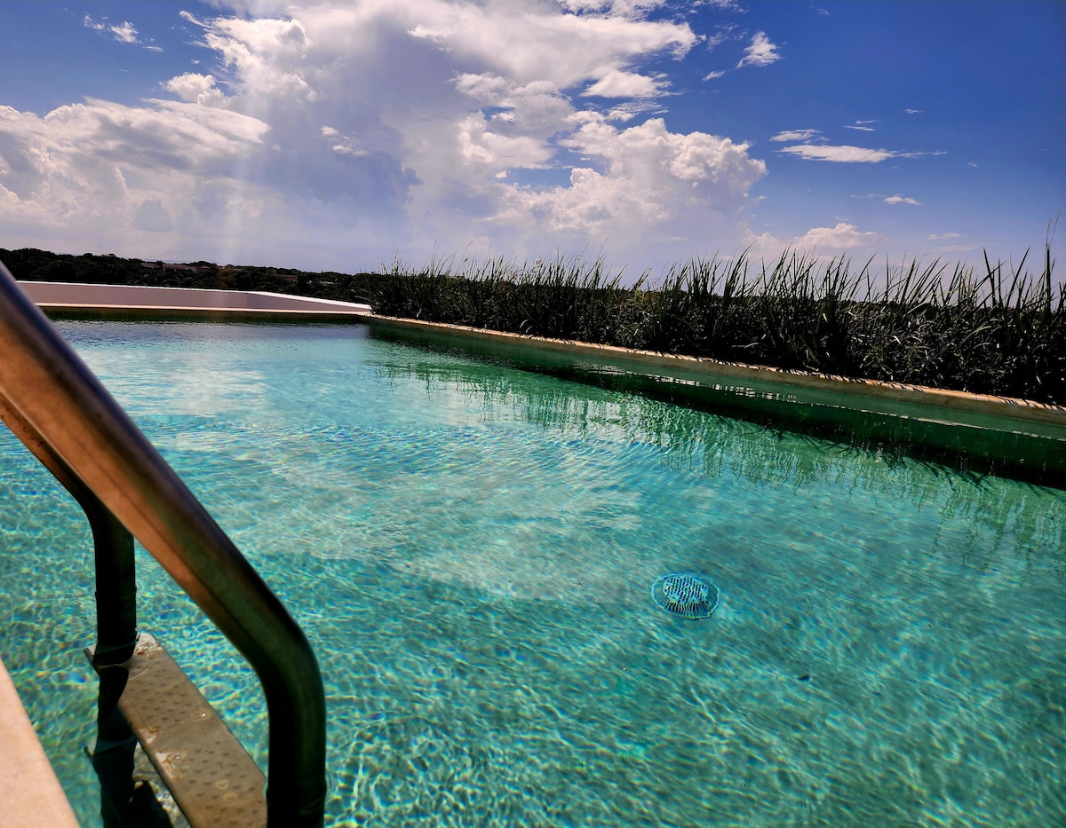 A clear swimming pool is highlighted by the reflection of clouds above. Steps lead from the edge of the pool toward the tranquil water, surrounded by lush greenery that contrasts against the blue sky.