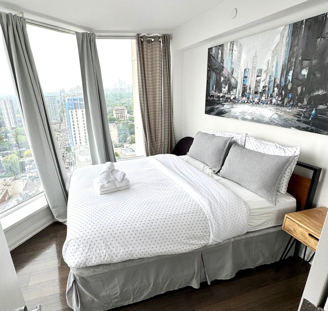A well-lit bedroom features a spacious bed dressed with neutral bedding and decorative pillows. A painting of an urban landscape adorns the wall above the bed. Large windows provide panoramic city views, while curtains softly frame the natural light.