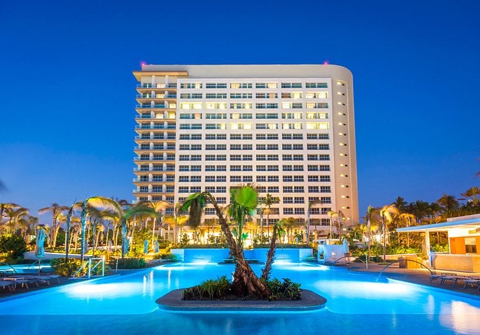 The exterior of a large resort building is illuminated at dusk, showcasing multiple floors and numerous windows. A tranquil pool area is surrounded by palm trees and lush landscaping, reflecting the warm glow of outdoor lighting.