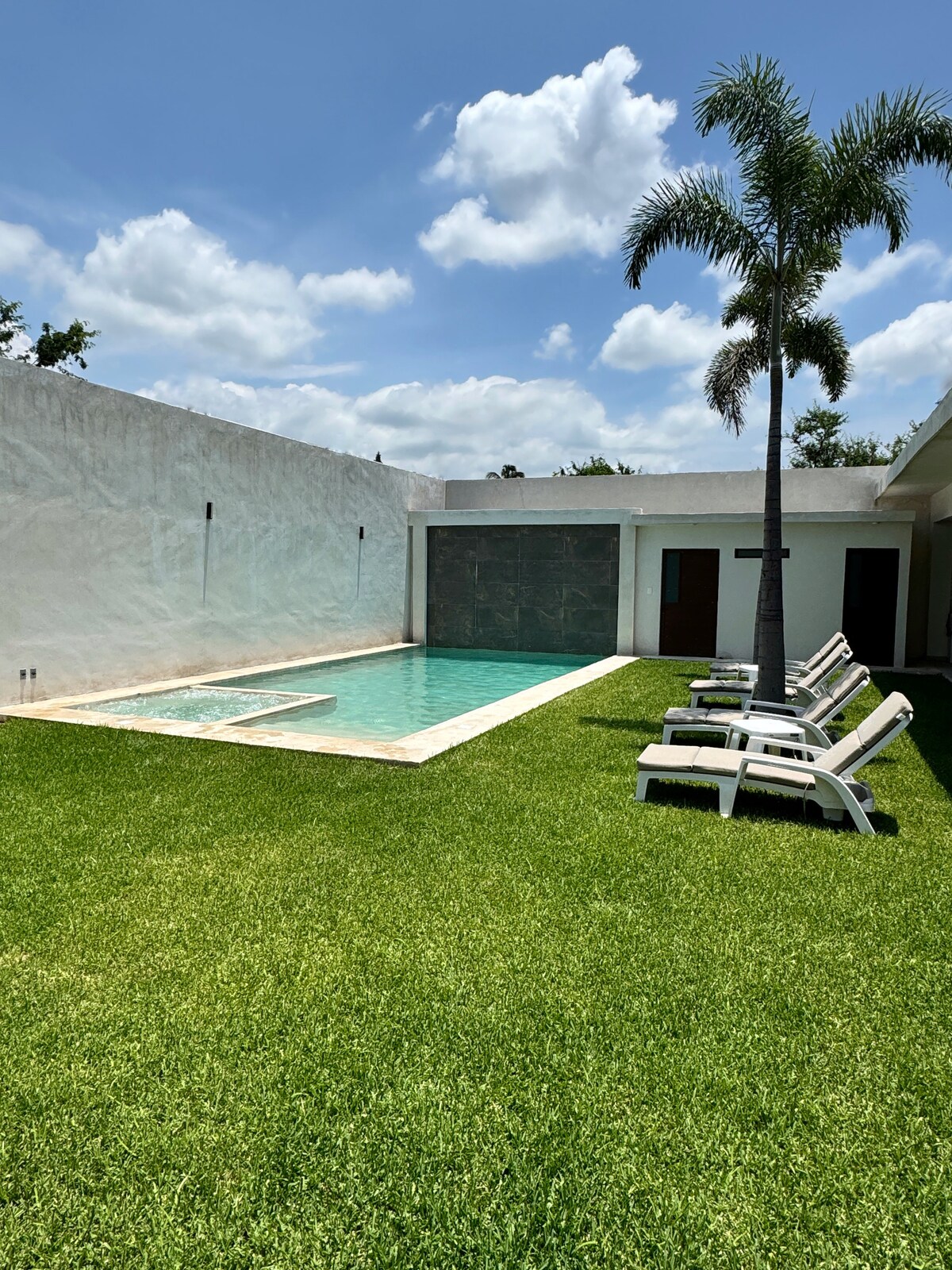 A private pool is surrounded by a neatly trimmed lawn and is complemented by a palm tree. Sun loungers line the poolside, offering a space for relaxation. The clean white walls reflect sunlight, creating an open atmosphere under a partly cloudy sky.