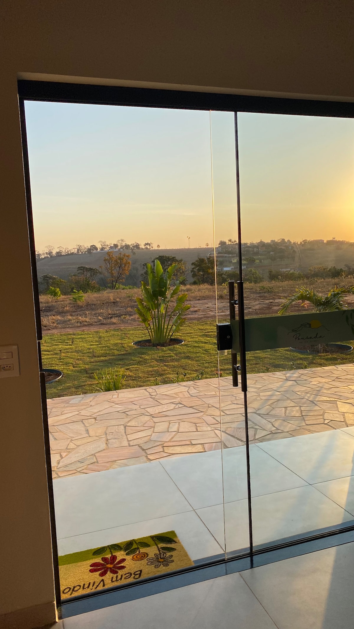A clear glass door stands open, revealing a scenic view of a grassy landscape bathed in warm sunlight. Lush greenery is visible in the foreground, complemented by a stone pathway leading outside. A colorful welcome mat is placed just inside the entrance.