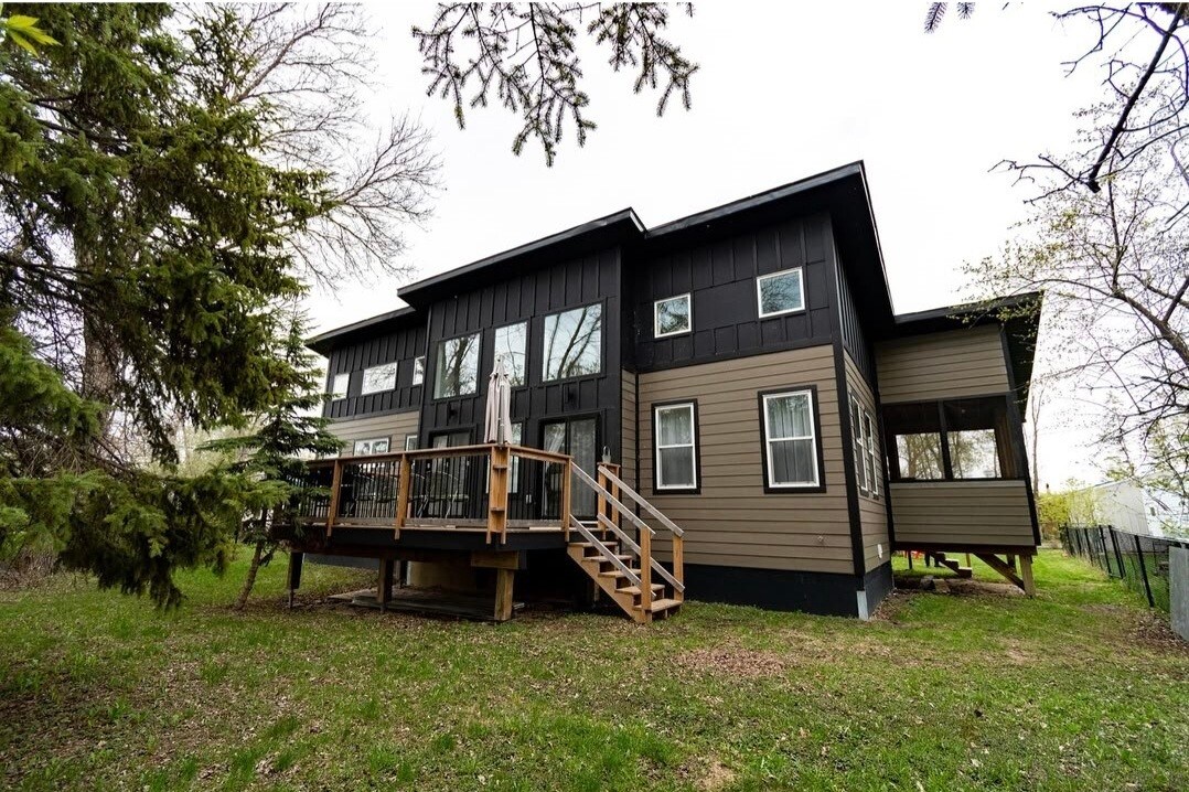 The two-story home features a mix of dark and light exterior siding, surrounded by greenery. A spacious deck is visible, providing access to outdoor seating. Large windows create a bright appearance, while the design incorporates multiple entry points under a canopy of trees.