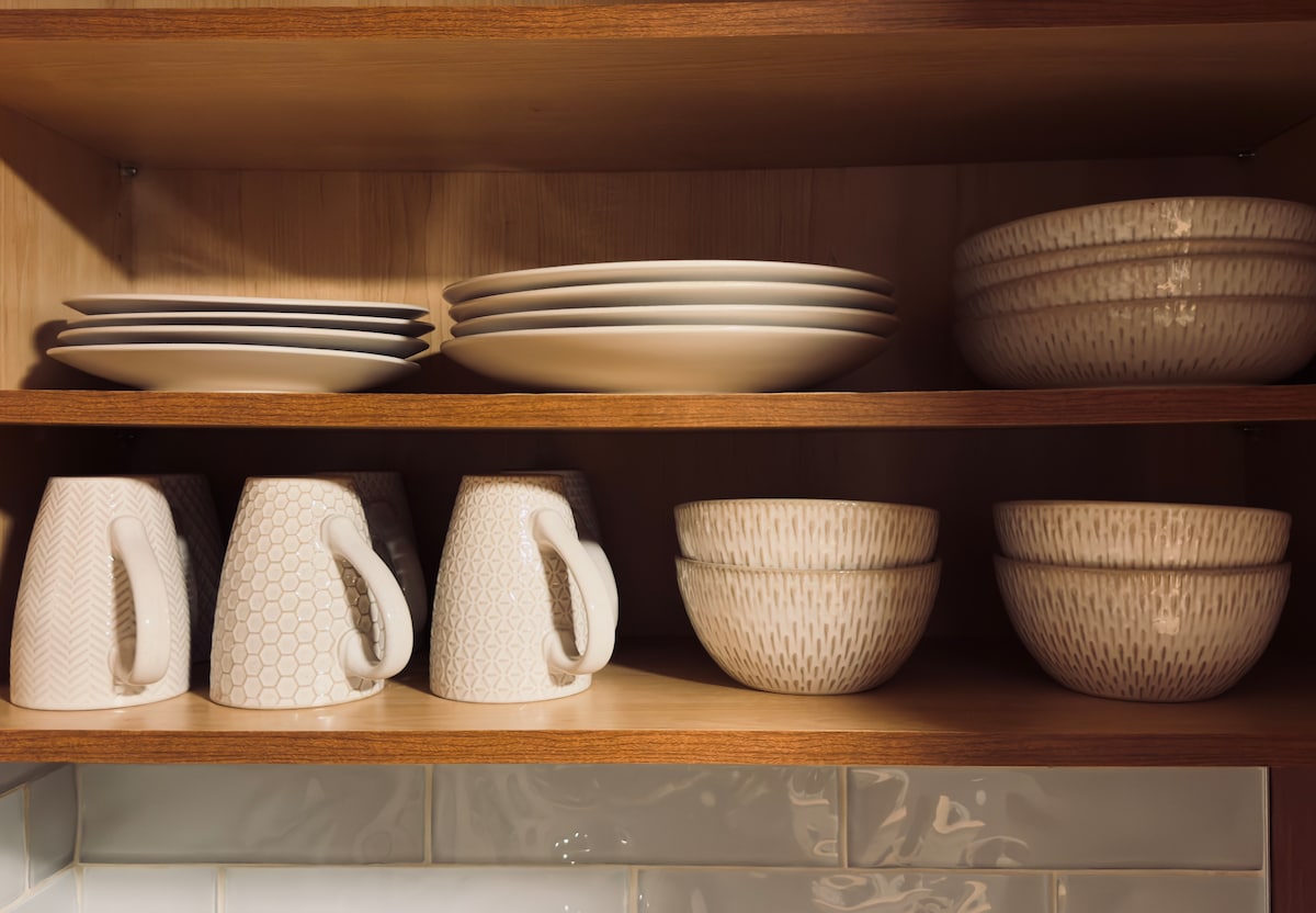 Neutral dishware is neatly arranged on wooden shelves. The collection includes several plates, cups, and bowls featuring subtle patterns, creating a cohesive and inviting display in the cottage's kitchen area.