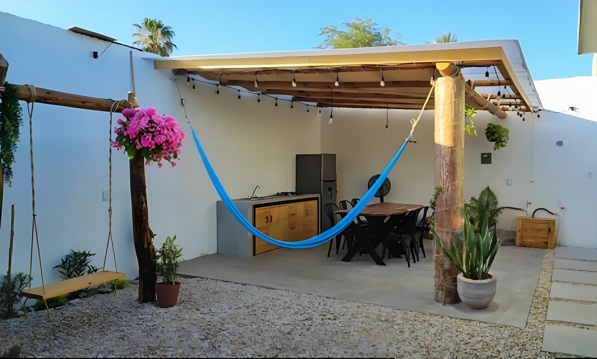 An outdoor patio features a hammock suspended between posts, alongside a dining table surrounded by black chairs. A shaded area is provided by a roof supported by wooden beams. Potted plants and vibrant pink flowers add a touch of greenery, enhancing the inviting space.