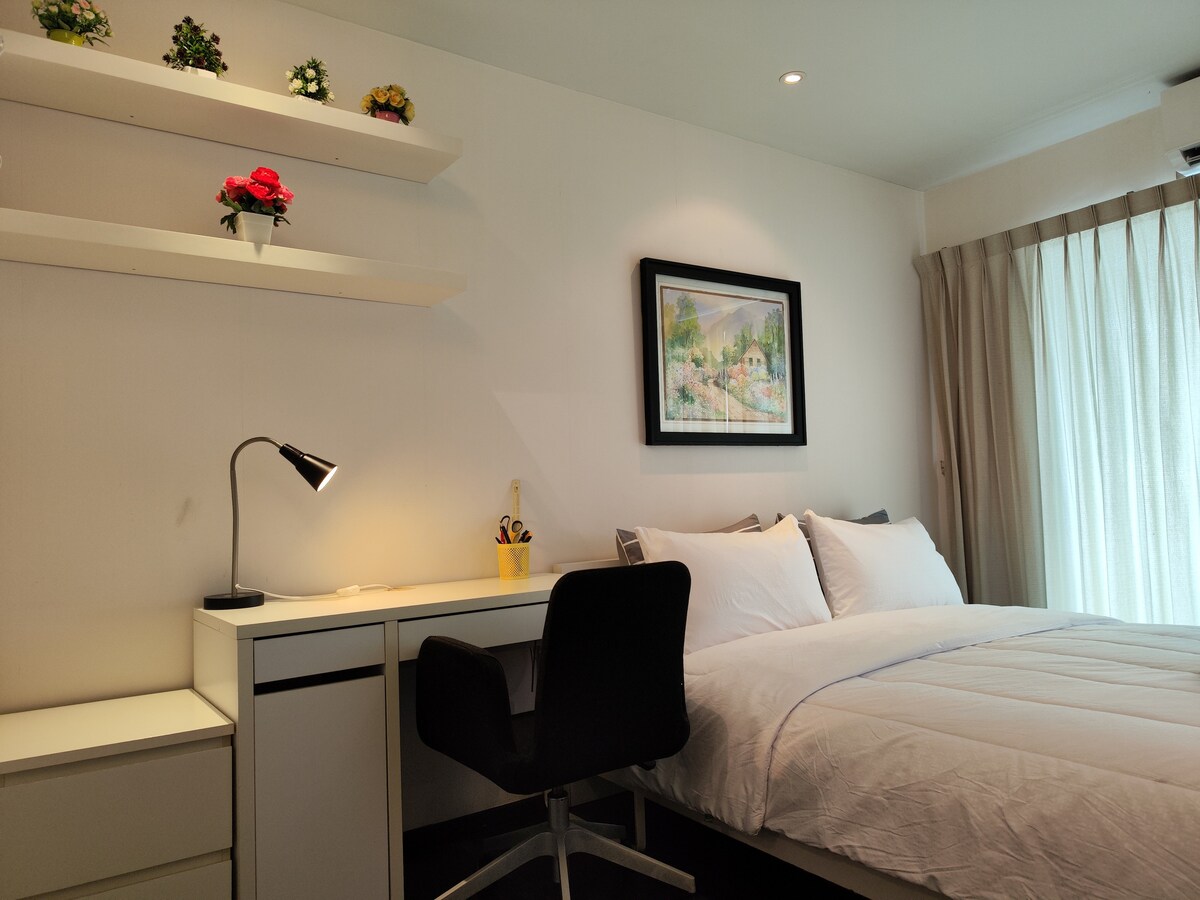 A studio room features a spacious bed with white linens, complemented by colorful decorative pillows. A desk workspace with a lamp is positioned beside the bed, and a shelf above displays small plants and a framed art piece. Soft natural light filters through sheer curtains.