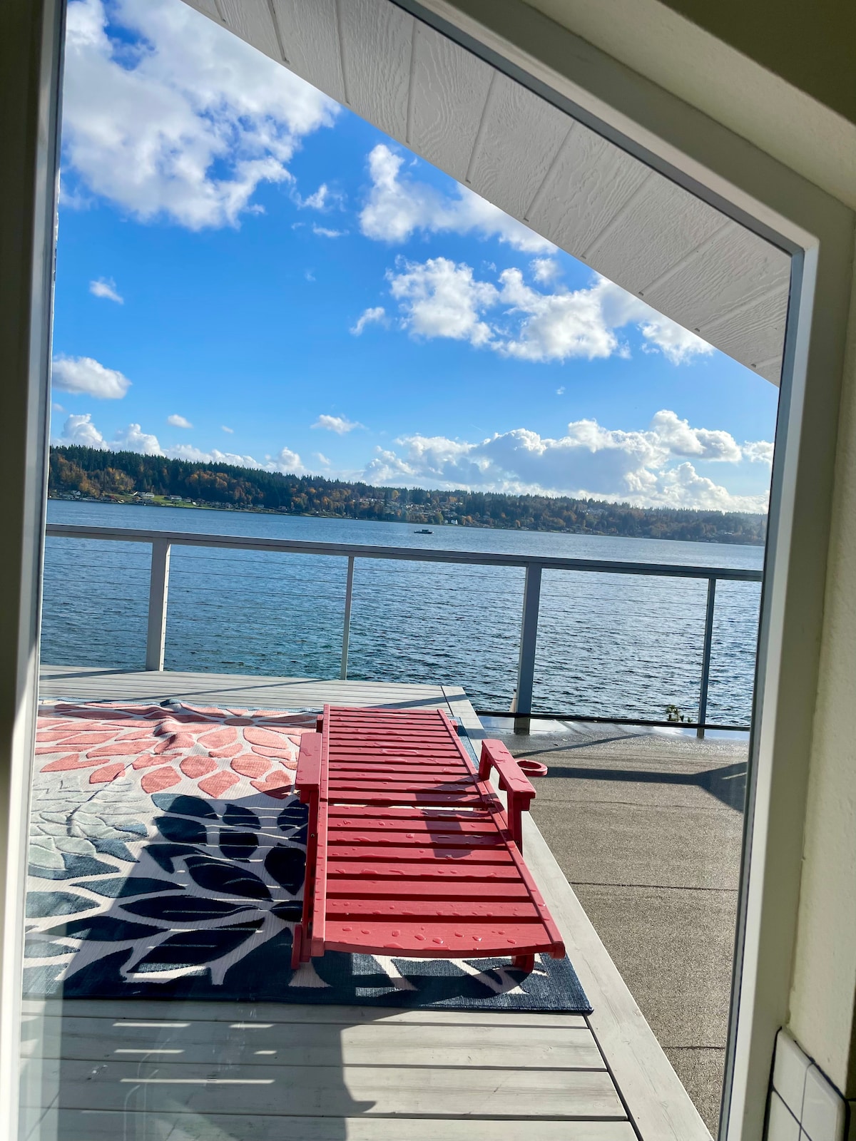 A view from inside the home frames a red lounge chair positioned on a deck overlooking the water. A patterned rug adds color beneath the chair, while the bright sky and surrounding landscape reflect a serene atmosphere.