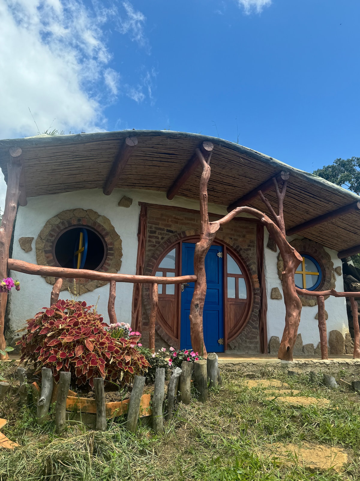 A charming hobbit house is set against a clear blue sky, featuring a round blue door framed by natural wood beams. Colorful flowers are planted around the entrance, with stone details highlighting the rustic design. The structure blends harmoniously with the surrounding greenery.