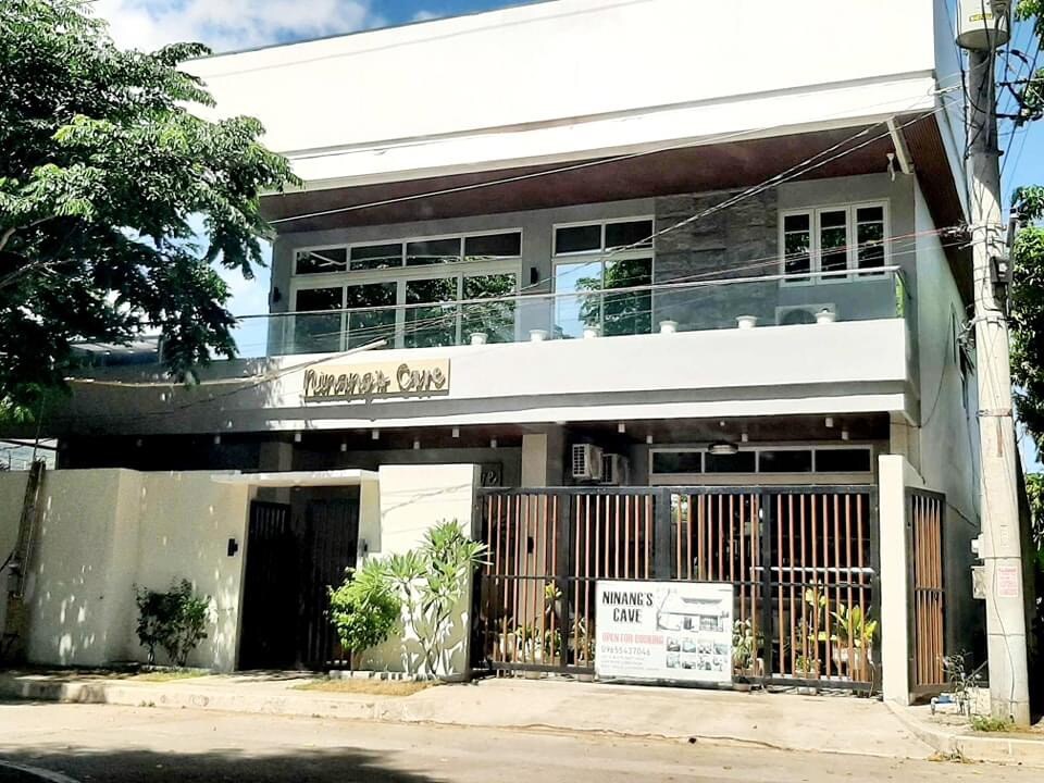 A modern two-story building with large glass windows, allowing natural light to fill the space. A welcoming entrance is framed by tropical plants and a wooden gate. The sign for Ninang’s Cave is displayed prominently above the entrance, indicating the resort's name.