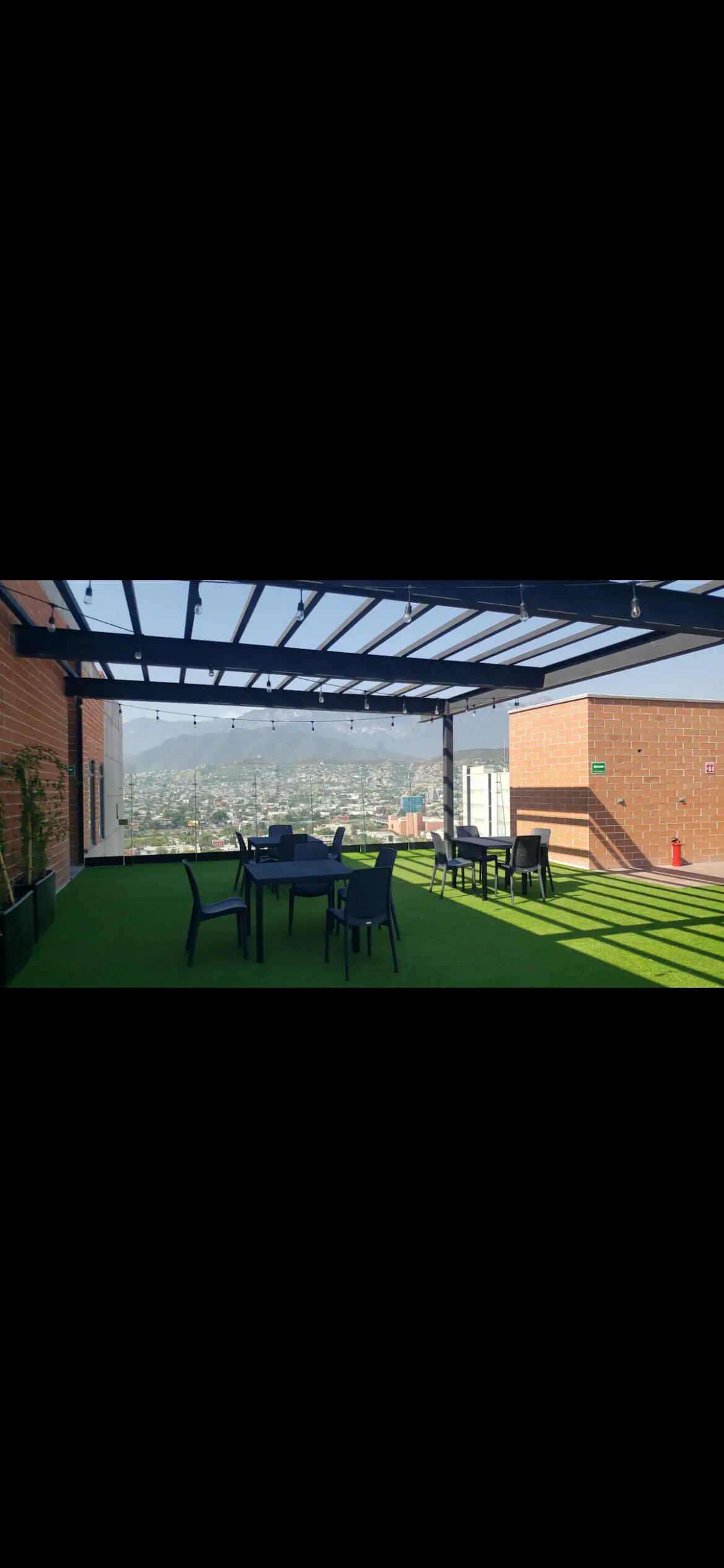 A spacious rooftop terrace features neatly arranged outdoor seating with tables and chairs. A shaded area is provided by a modern pergola, and stunning views of the surrounding mountains are visible in the background. Lush greenery adds a touch of nature to the setting.
