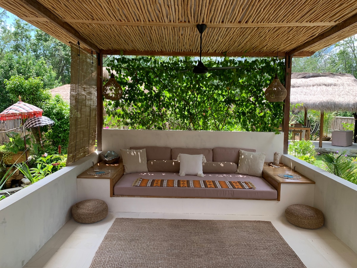 An inviting outdoor lounge area features a large cushioned bench adorned with decorative pillows, set against a backdrop of lush greenery. Natural materials such as bamboo cover the ceiling, while earthy tones are prominent in the furnishings, creating a relaxed atmosphere.
