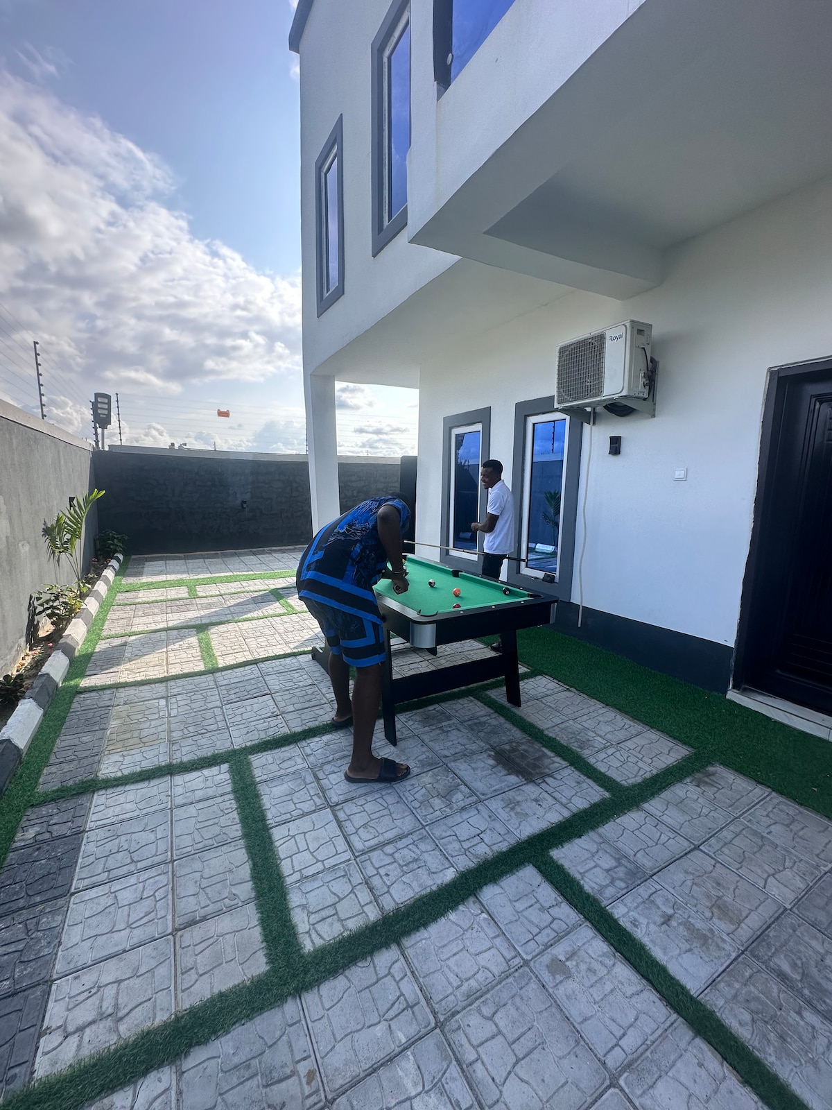 An outdoor space features a snooker table, set on a grassy pathway lined with decorative stones. The area is illuminated by natural light, with a modern building structure in the background. A person can be seen positioned at the snooker table, engaged in play.
