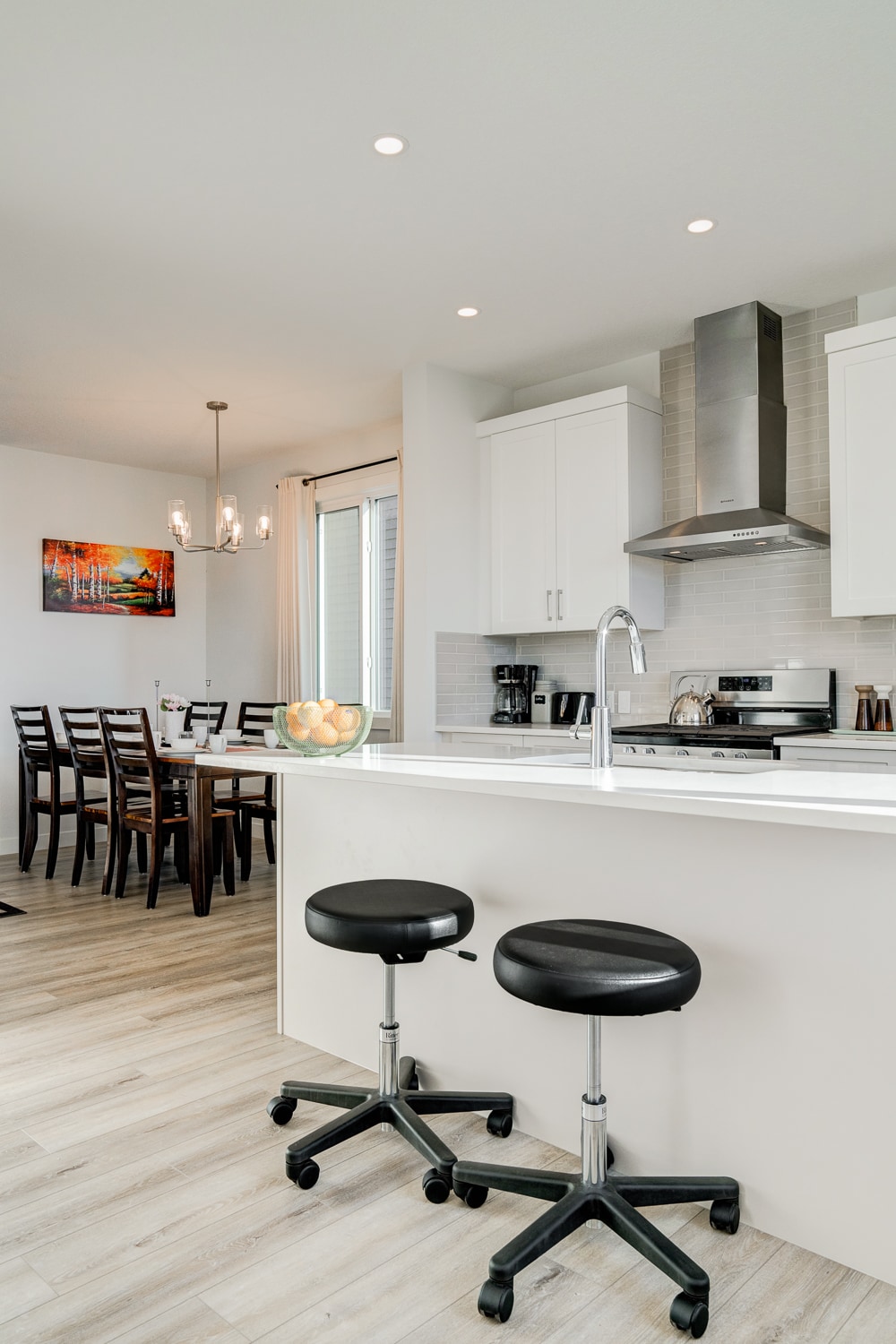 A modern kitchen is featured with white cabinetry and stainless steel appliances. Two adjustable black stools are placed at the island. A dining area with a wooden table and six chairs is visible in the background, illuminated by natural light through sliding glass doors.
