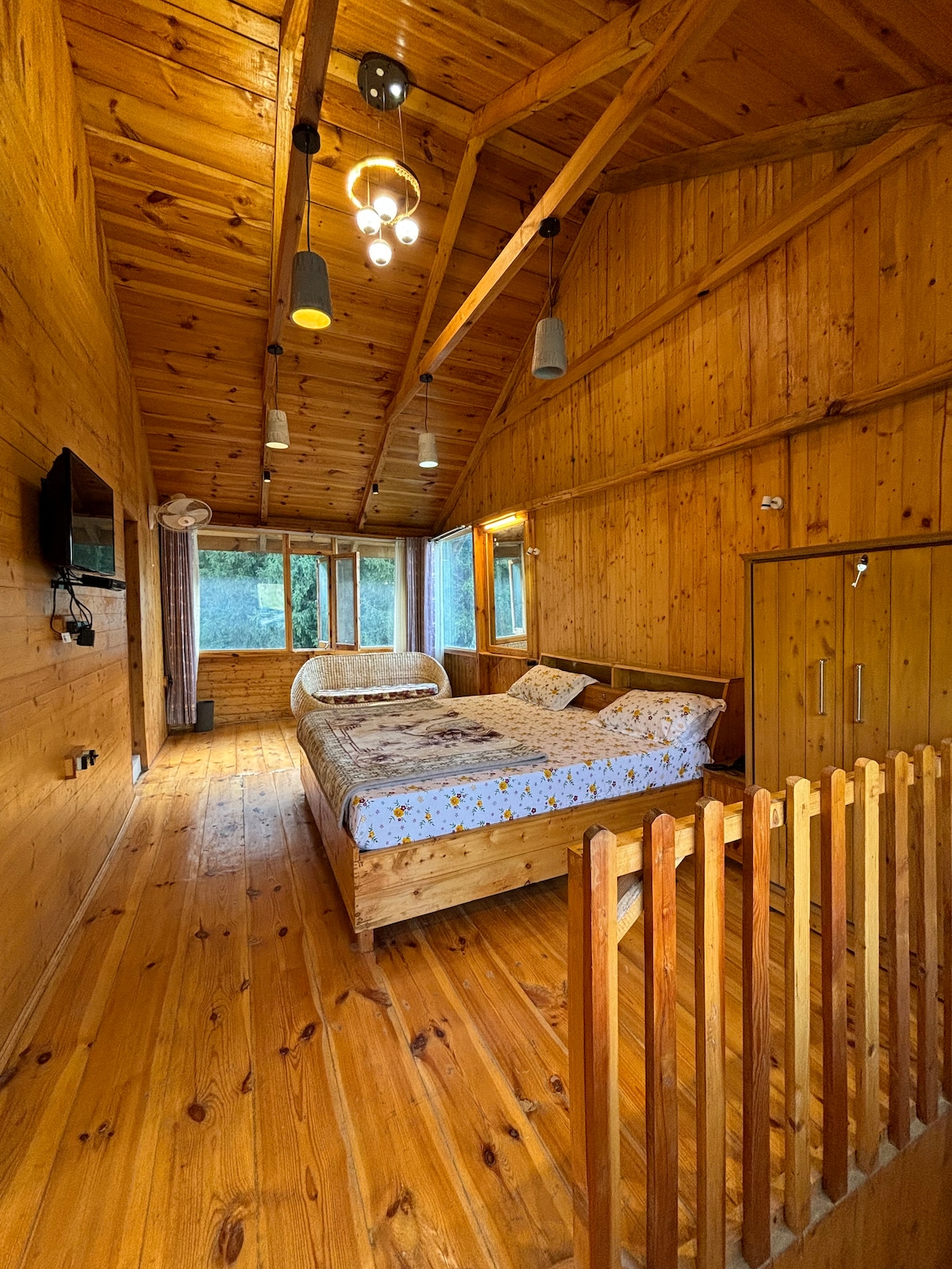 A wooden interior creates a warm ambiance in this spacious bedroom. Natural light streams through large windows, illuminating a generous bed arranged with patterned linens. A rustic railing divides the sleeping area from the open space, while a flat-screen TV is mounted on the wall.