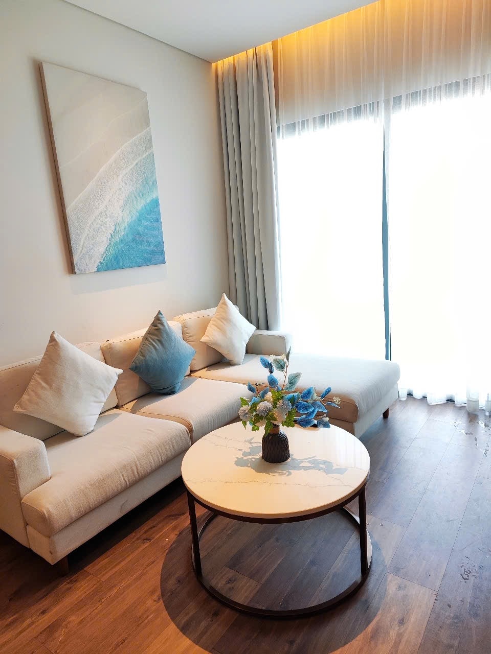 A light-filled living area features a soft sectional sofa adorned with decorative cushions. A small round coffee table rests in the center, complemented by a simple vase with blue flowers. Floor-to-ceiling windows allow glimpses of natural light and outdoor views.
