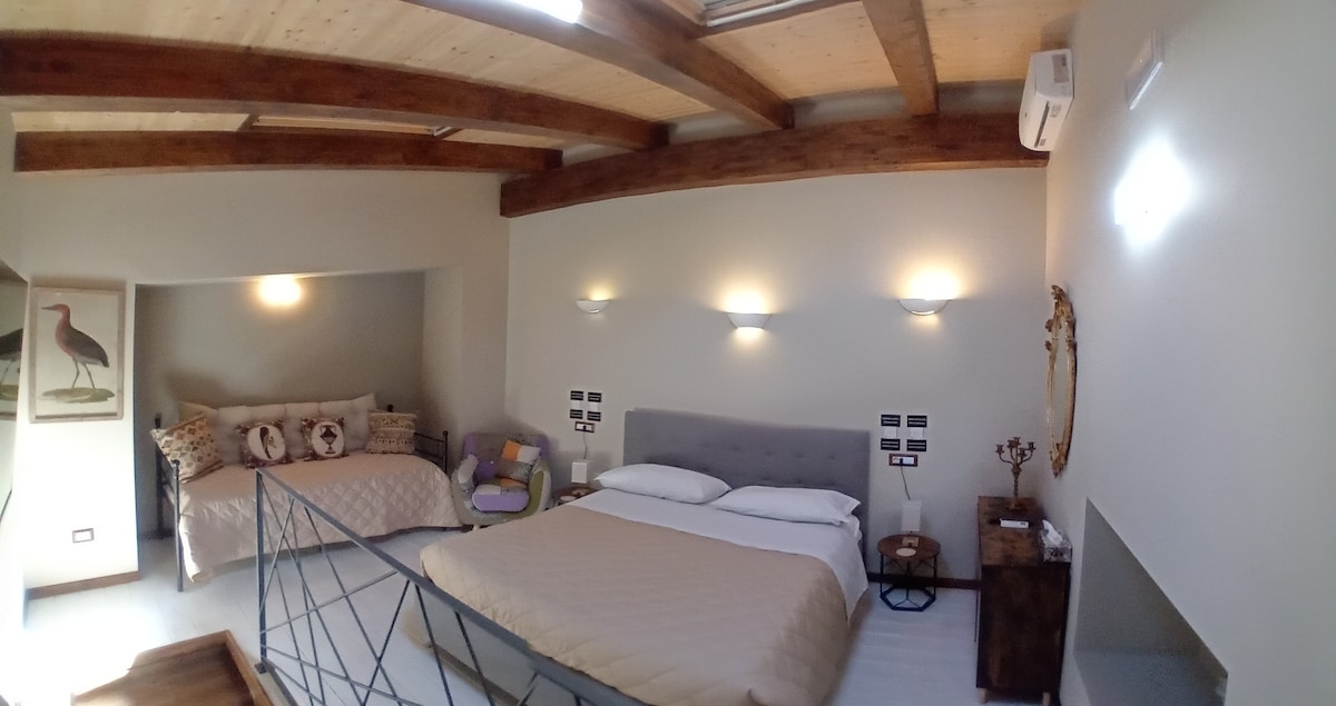 A spacious bedroom features a comfortable bed with soft linens, accompanied by a cozy daybed nearby. Neutral walls are complemented by wooden beams overhead, providing warmth. Two wall sconces offer gentle lighting, while a decorative mirror enhances the inviting ambiance of the room.