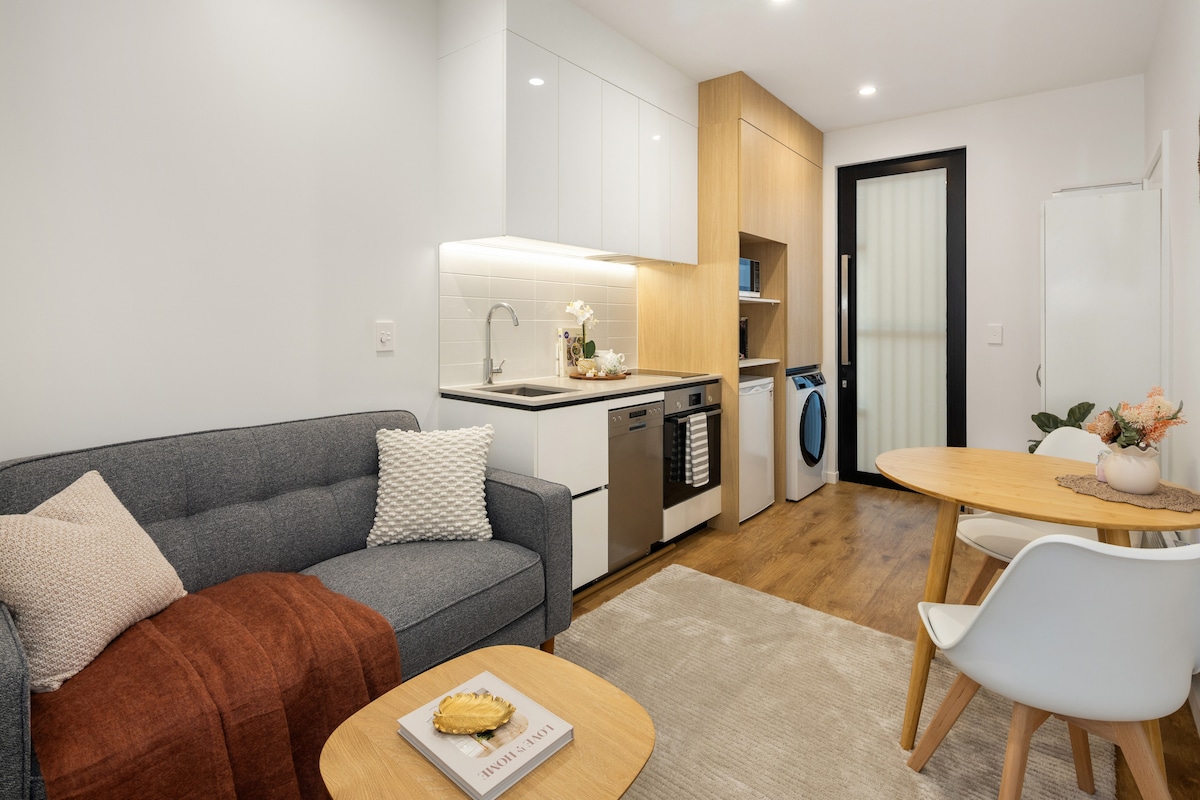 An open-plan living area features a comfortable sofa adorned with cushions and a rust-colored throw. A wooden dining table with white chairs is positioned nearby. The modern kitchen showcases sleek cabinetry, a sink, and essential appliances, including a refrigerator and washing machine.