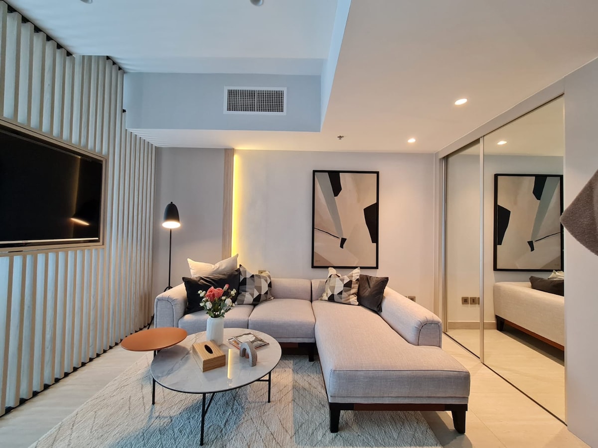 A modern living area is featured with a sectional sofa decorated with contrasting pillows. A round coffee table sits in front, accompanied by a vase of flowers. A large mirror reflects the room, while abstract artwork enhances the stylish ambiance.