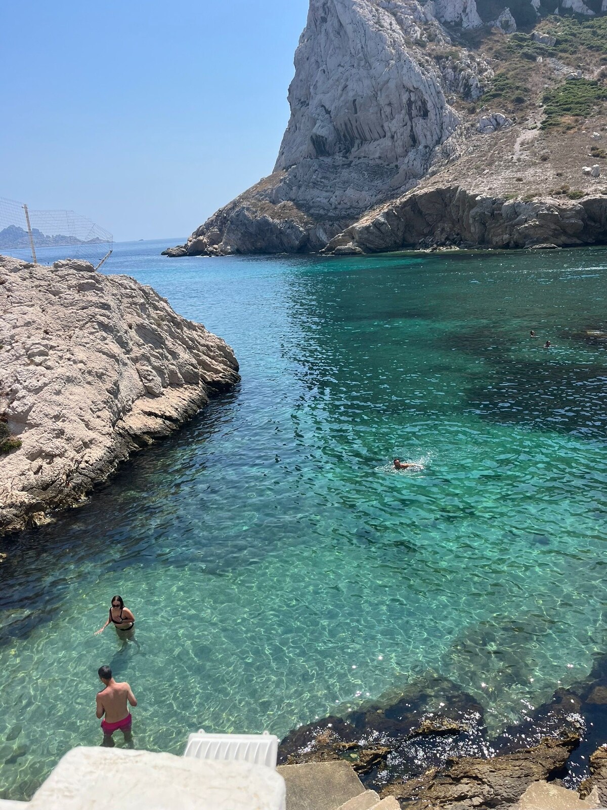 Vibrant turquoise waters are visible, reflecting sunlight over the rocky shoreline. A small group of people can be seen enjoying the water, with one individual swimming further out. A steep rocky cliff rises in the background, complementing the natural beauty of the coastal area.