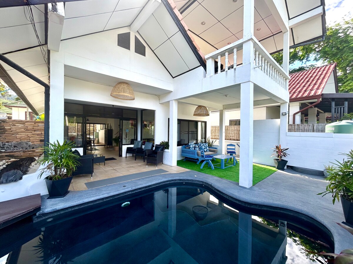 An outdoor area featuring a private pool surrounded by tropical plants. The space is complemented by modern seating arrangements, including lounge chairs and a casual dining area. The home’s architecture includes high ceilings and large glass doors that provide a view of the living space.