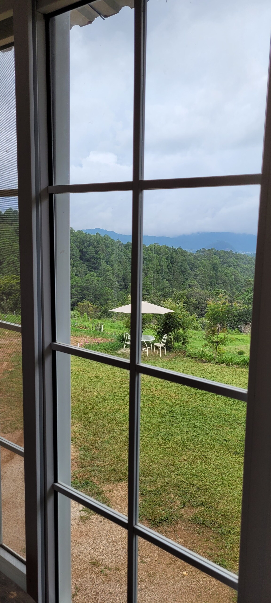 A view from a large window captures the green mountains beyond, with a small outdoor seating area visible in the foreground. The cloudy sky reflects a serene atmosphere, complementing the lush landscape.